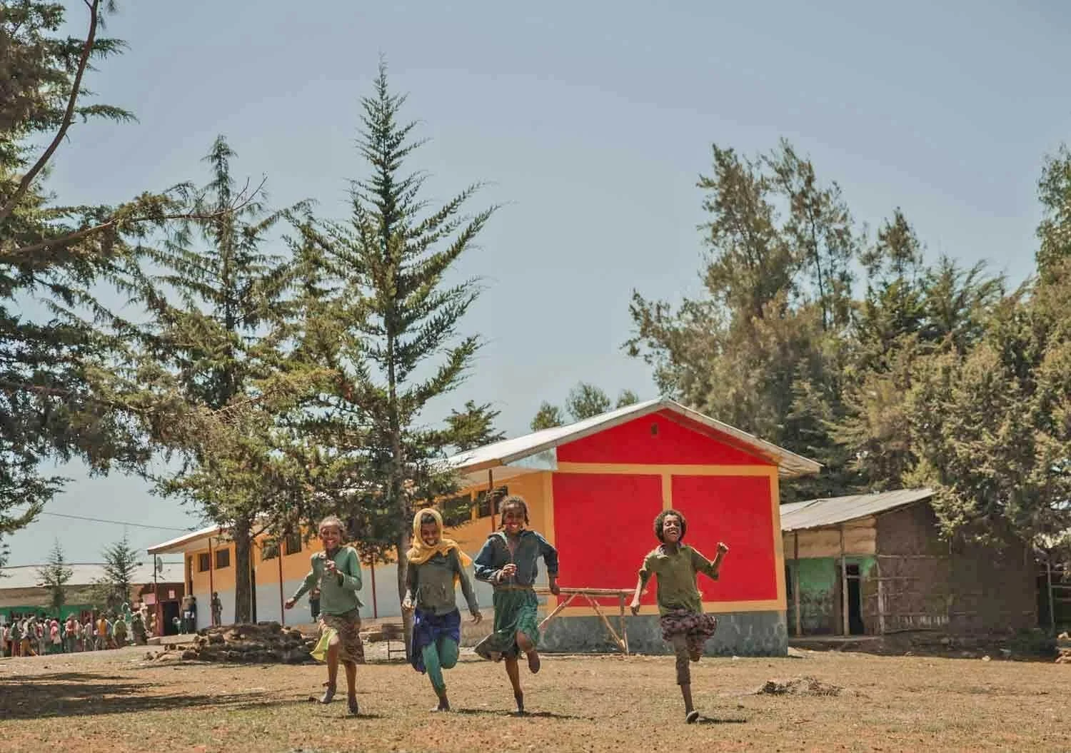 Four children running and playing on a dirt field in front of a red and yellow building surrounded by trees.