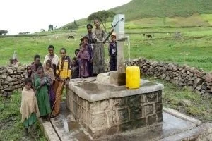 Group of children and a woman gather around a water well in a rural area with green hills in the background.