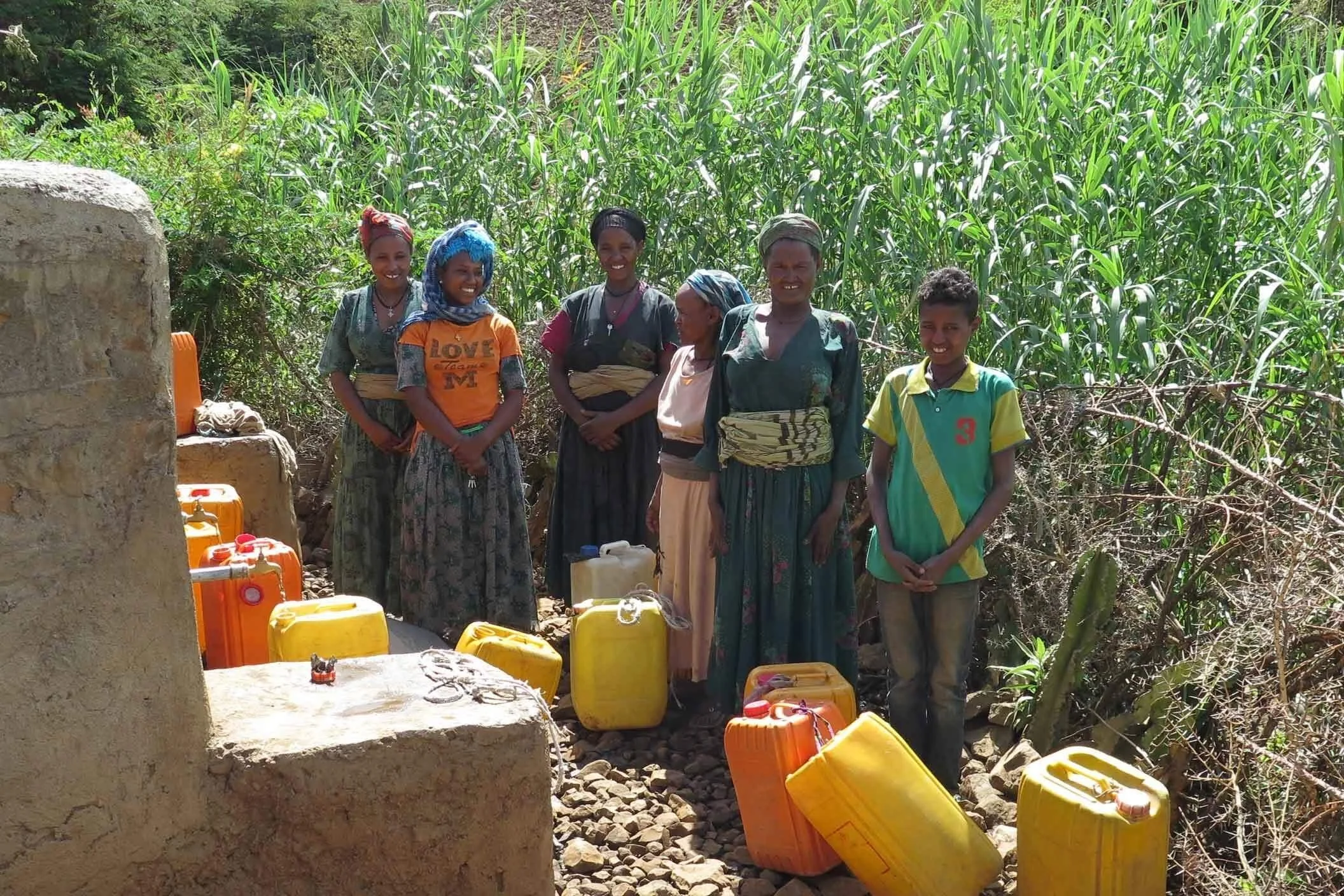 A group of seven people, including children and women, standing outdoors in front of tall green crops, with yellow and orange containers on the ground nearby.