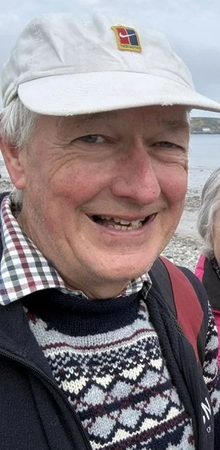 A smiling older man wearing a white baseball cap with a small patch, a checkered shirt, a patterned sweater, and a black jacket. He's outdoors near water.