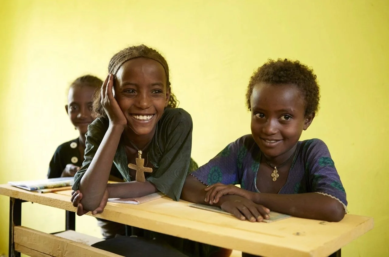 Three smiling children sitting at desks in a classroom with a yellow wall background.