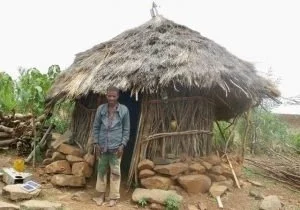 Man standing outside a small hut with a thatched roof, made of natural materials, in a rural area.
