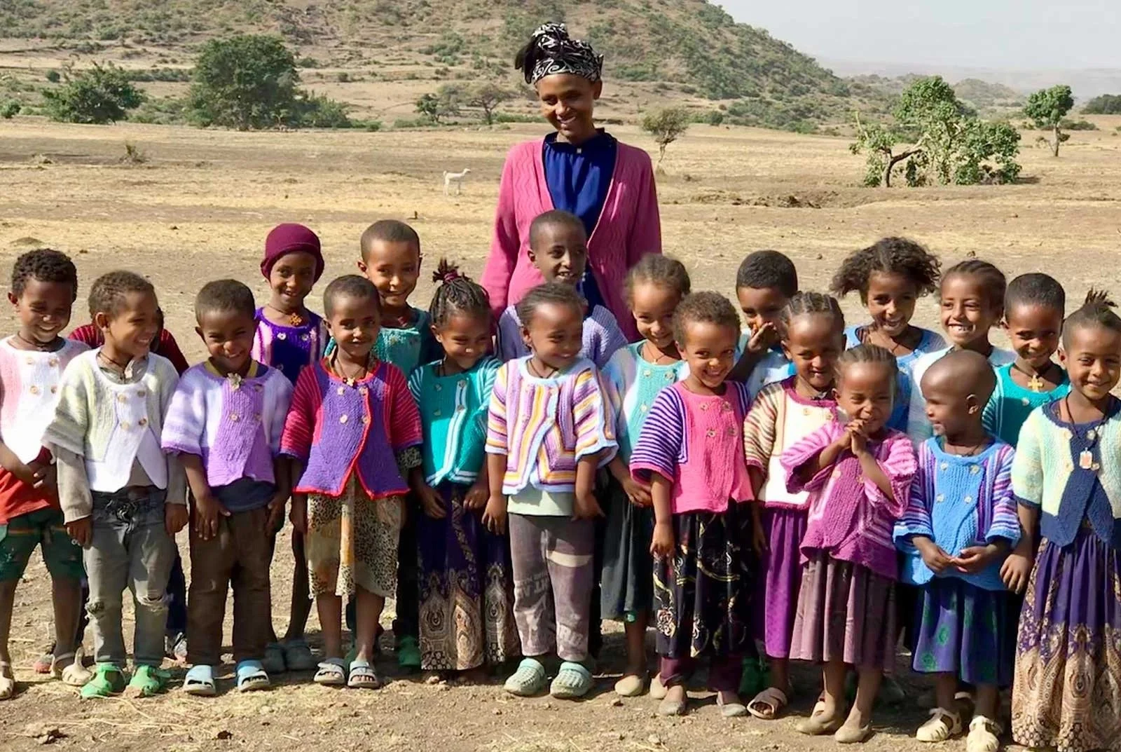 A group of young children, mostly girls, standing outdoors on a dirt ground with a woman behind them. The children are wearing colorful clothing and smiling, with some holding their hands together or at their sides. The landscape in the background shows a dry, hilly area with sparse trees and a mountain in the distance.