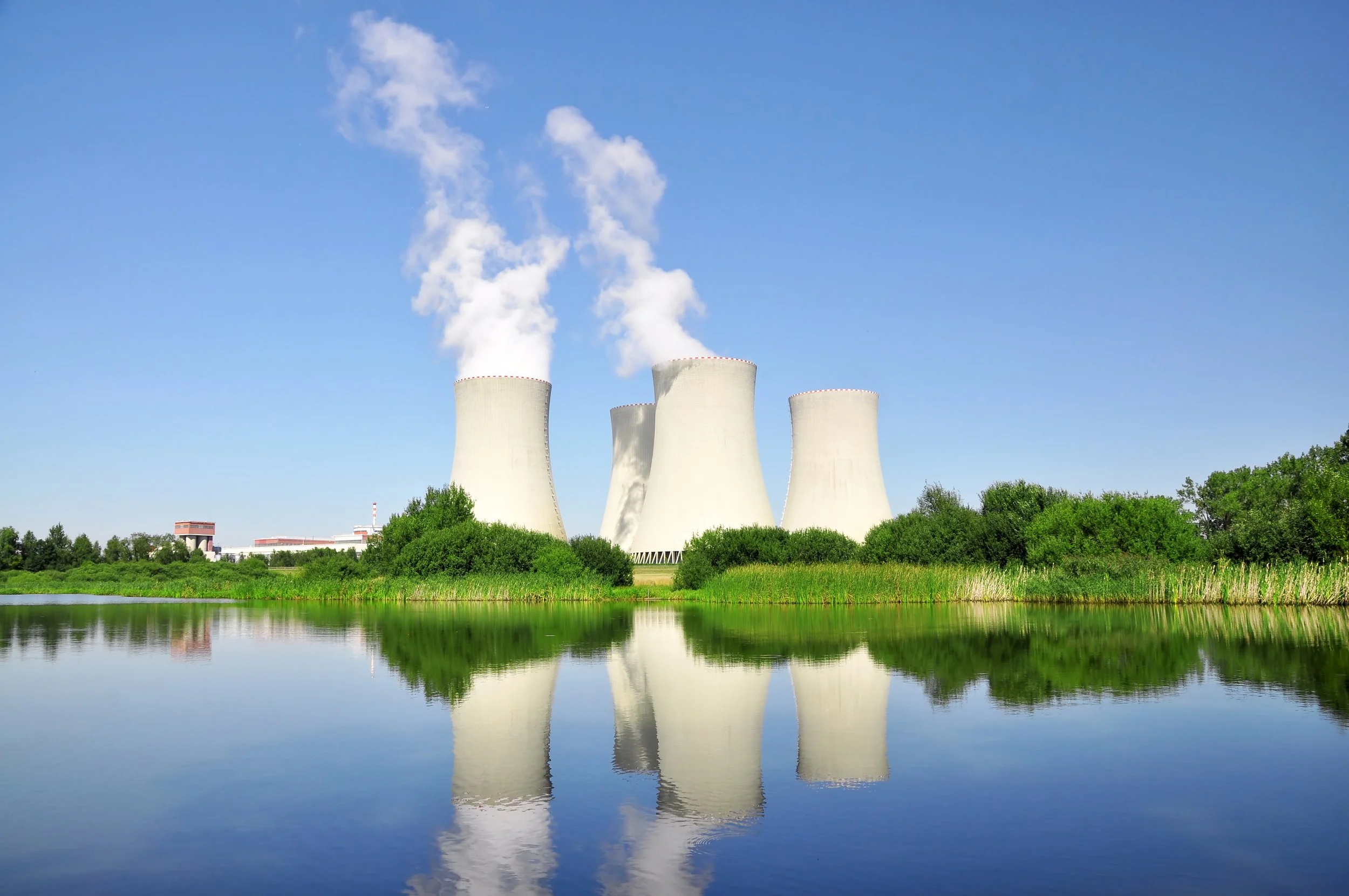Nuclear power plant with four cooling towers releasing steam into a blue sky, reflected in a body of water surrounded by green trees.