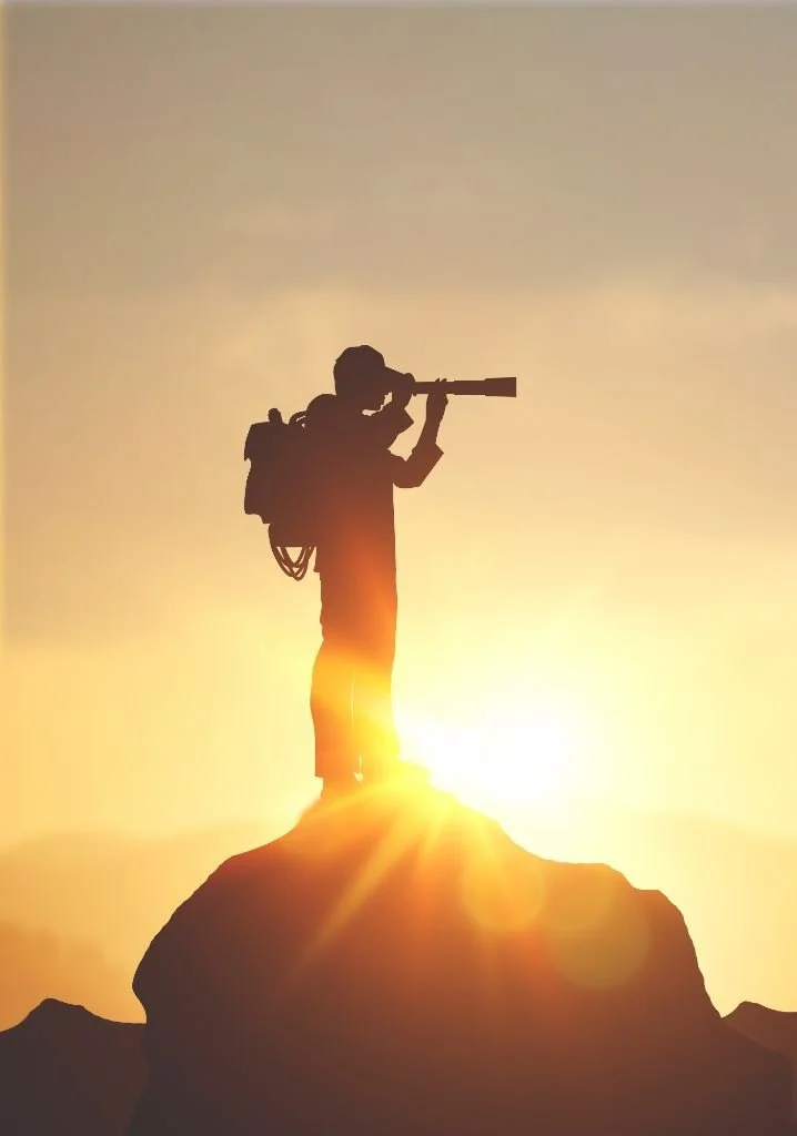 Silhouette of a person with a backpack using a telescope on a mountain at sunset.