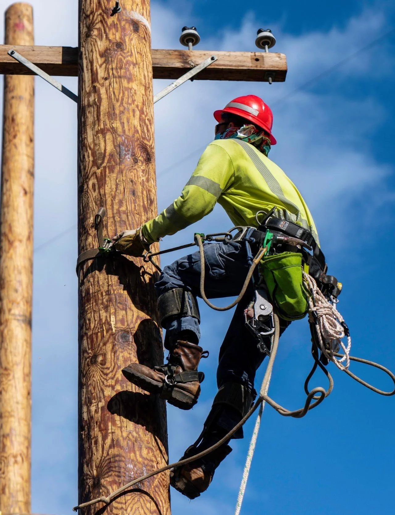 A lineman working on a wooden utility pole wearing safety gear including a red helmet, yellow high-visibility shirt, harness, gloves, and safety footwear, with power lines and equipment visible.