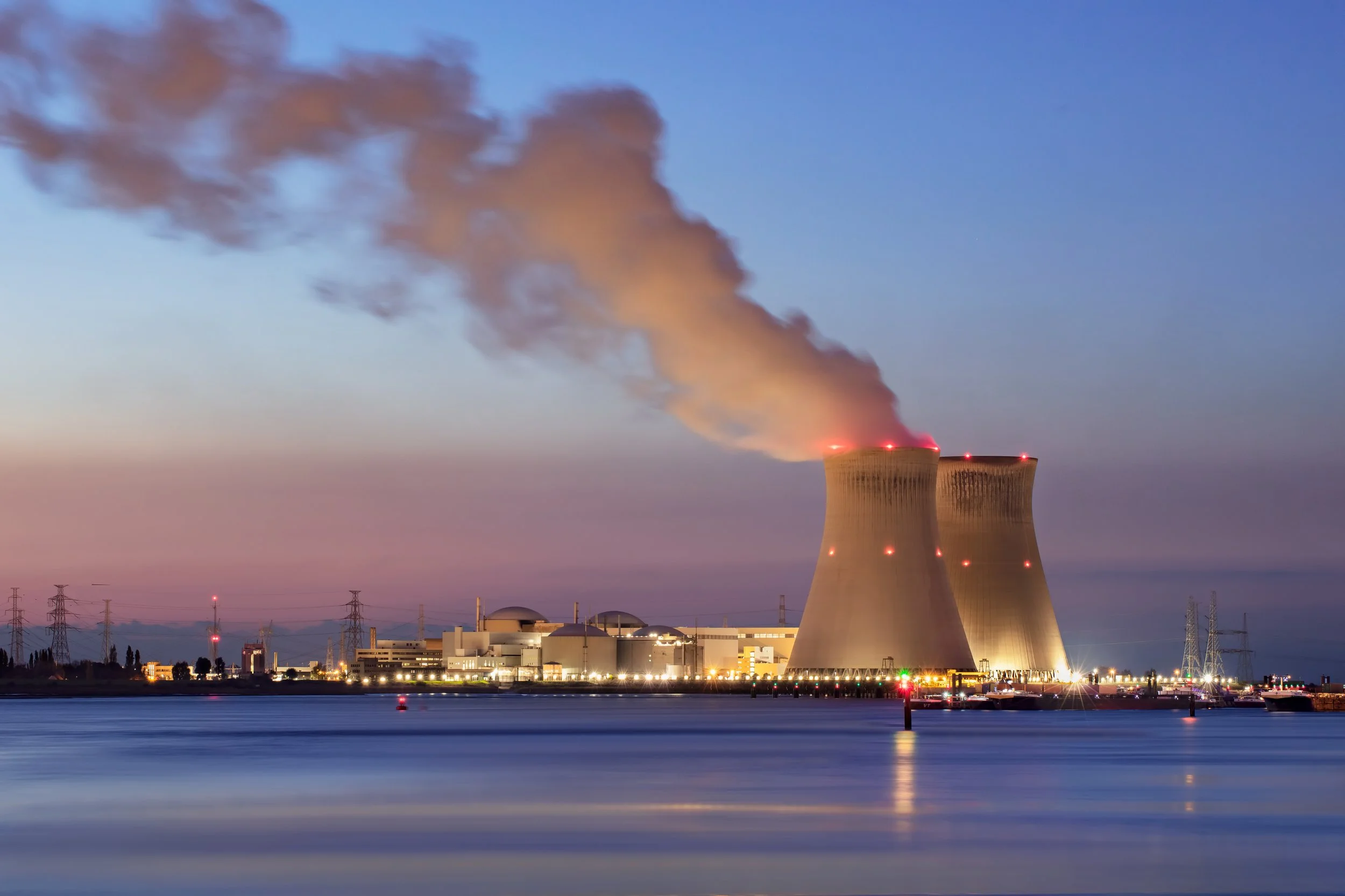 Cooling towers at a nuclear power plant releasing steam into the evening sky with city buildings and power lines in the background.