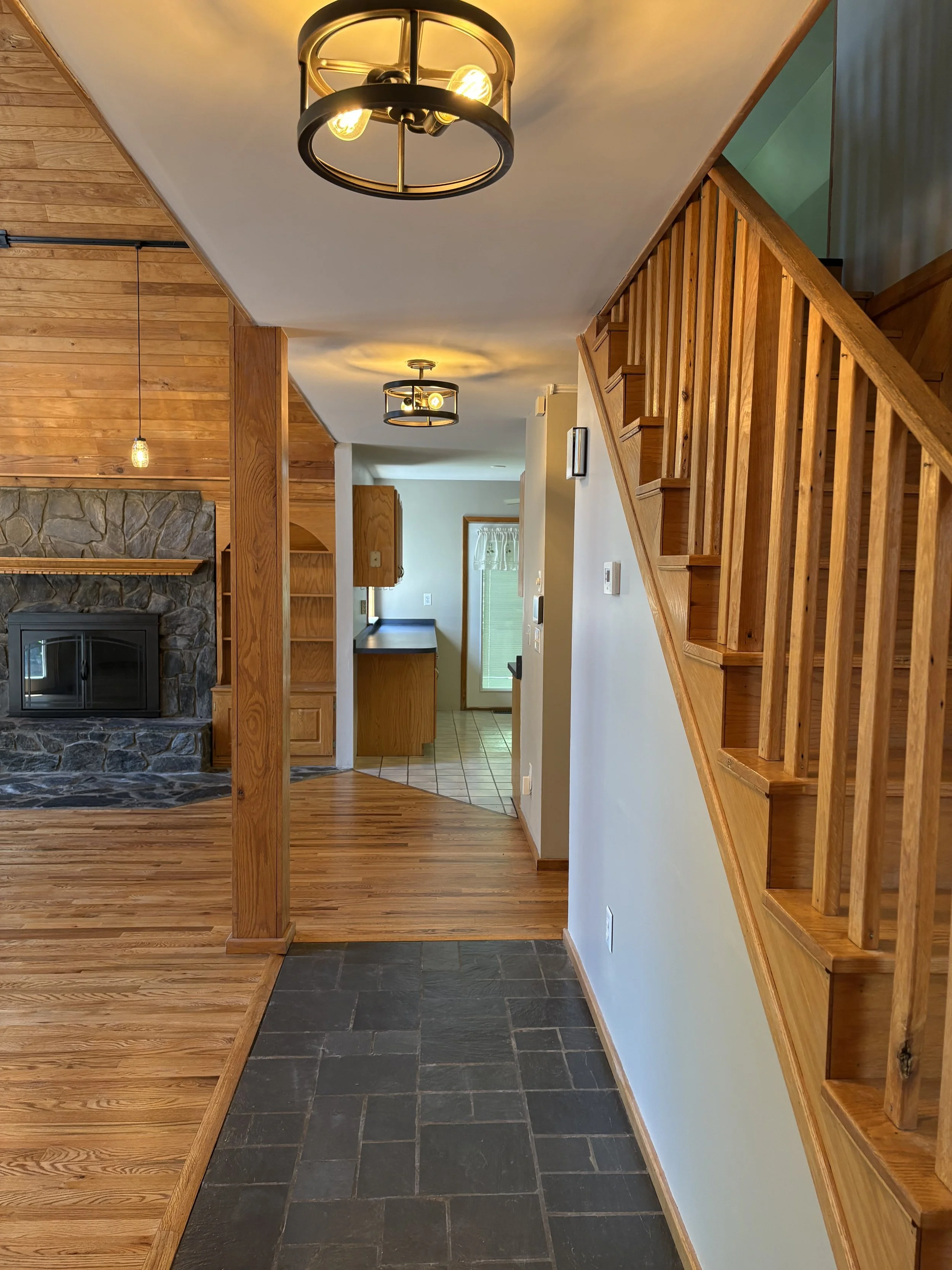 Interior view of a house with wooden flooring, a staircase with wooden banisters, a stone fireplace, and ceiling lights. A small foyer area with tile flooring is also visible.