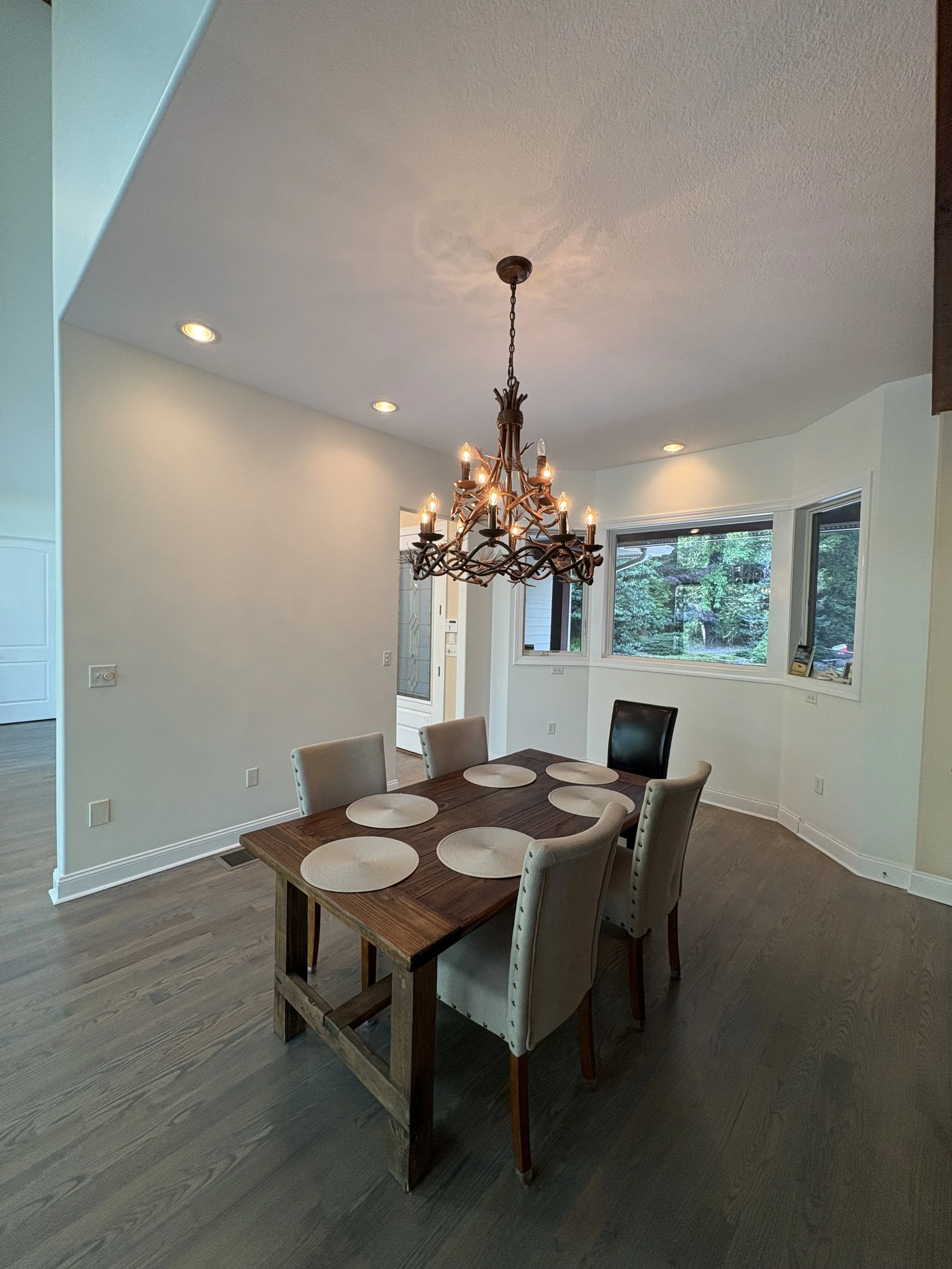 A dining room with a wooden table, beige upholstered chairs, a rustic chandelier, and large windows showing greenery outside.
