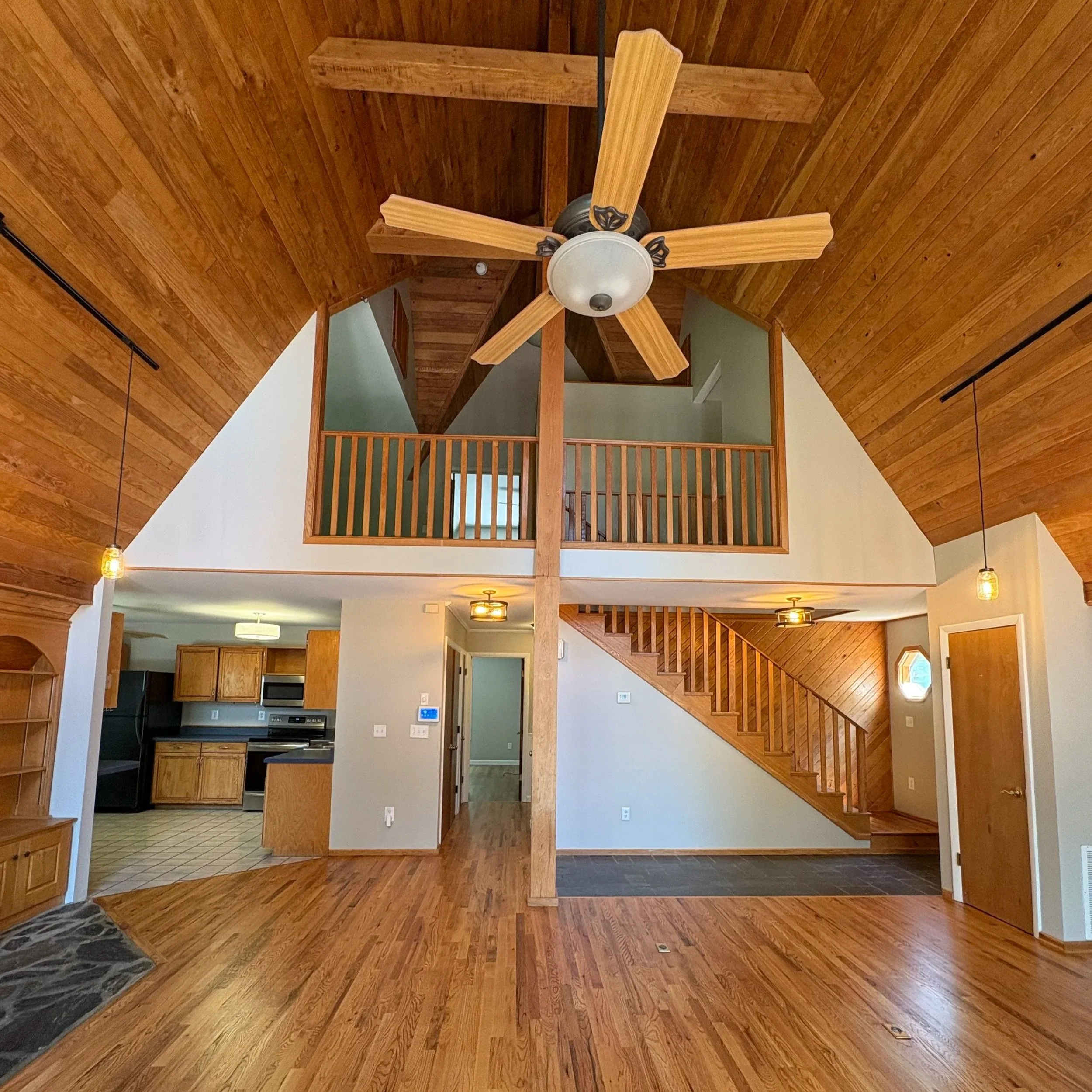 Interior view of a house with wooden floors, walls, and ceiling, showing a staircase with wooden railing and several hanging light fixtures.