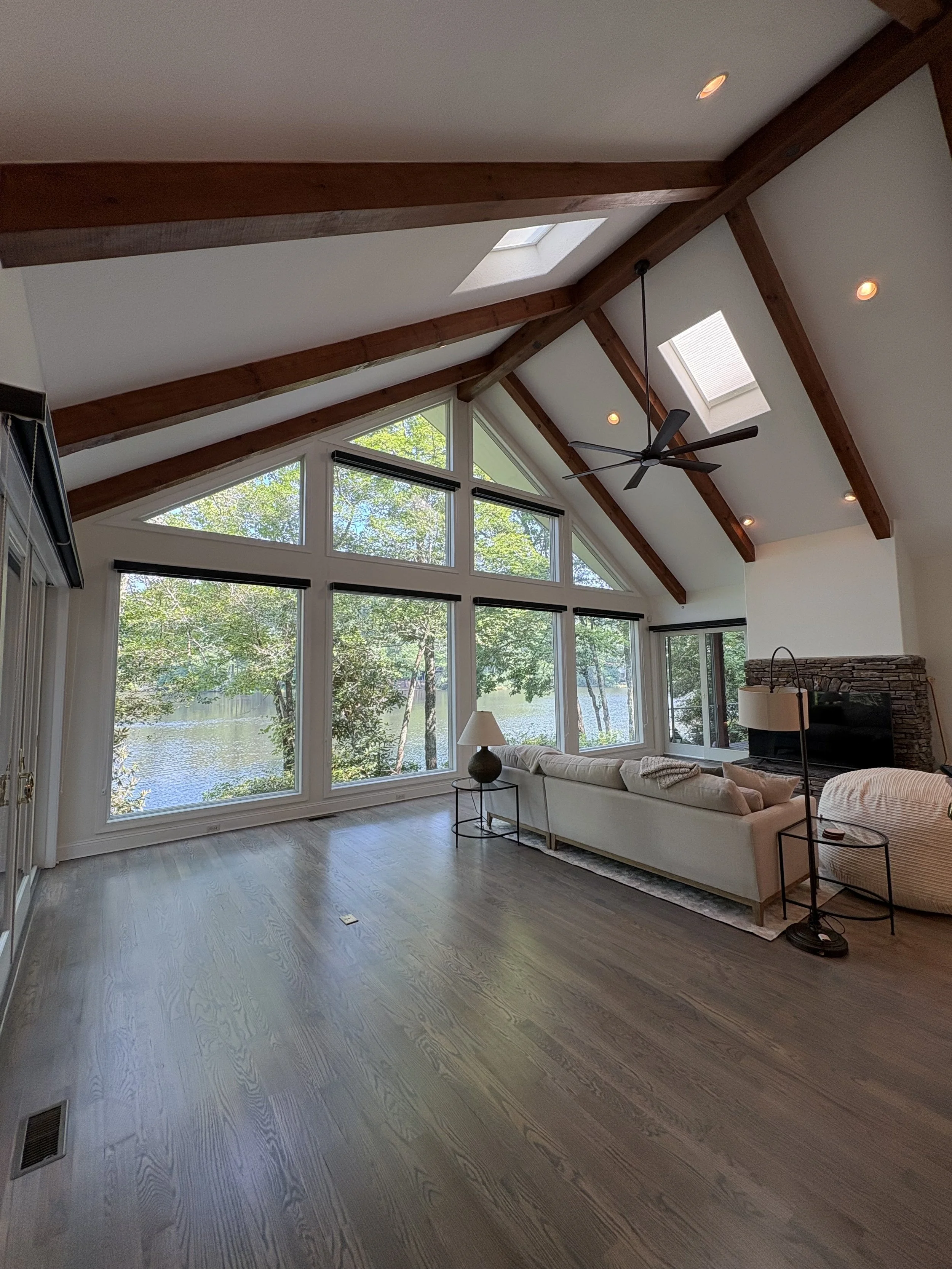 A spacious living room with large windows overlooking a wooded area and a lake. The ceiling features exposed wooden beams, skylights, and a ceiling fan. There is a cream-colored sofa, a side table with a lamp, and a stone fireplace.