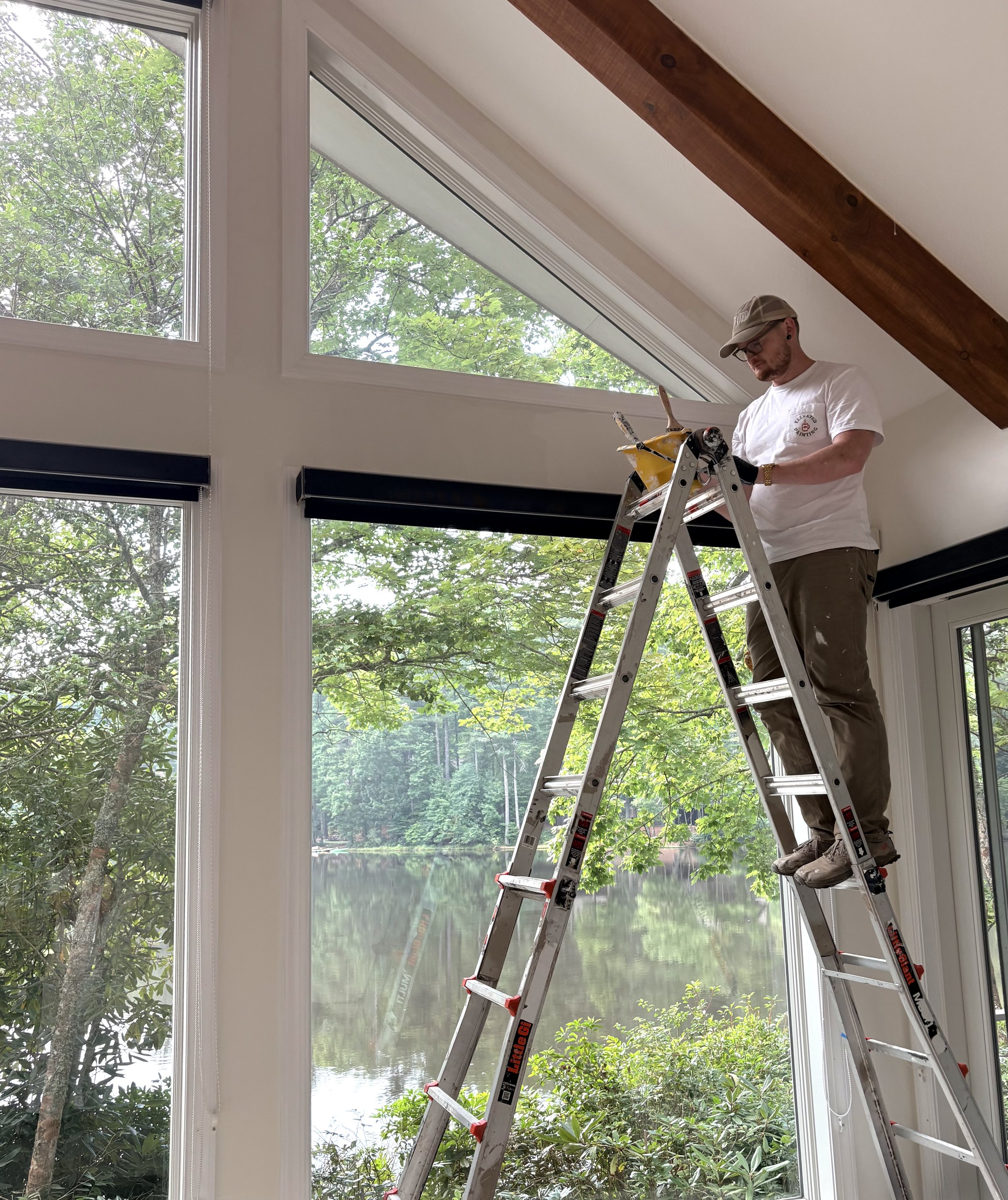A man standing on a ladder inside a room near large windows, looking down at his phone. Outside the windows, there is greenery with trees and a body of water in the background.