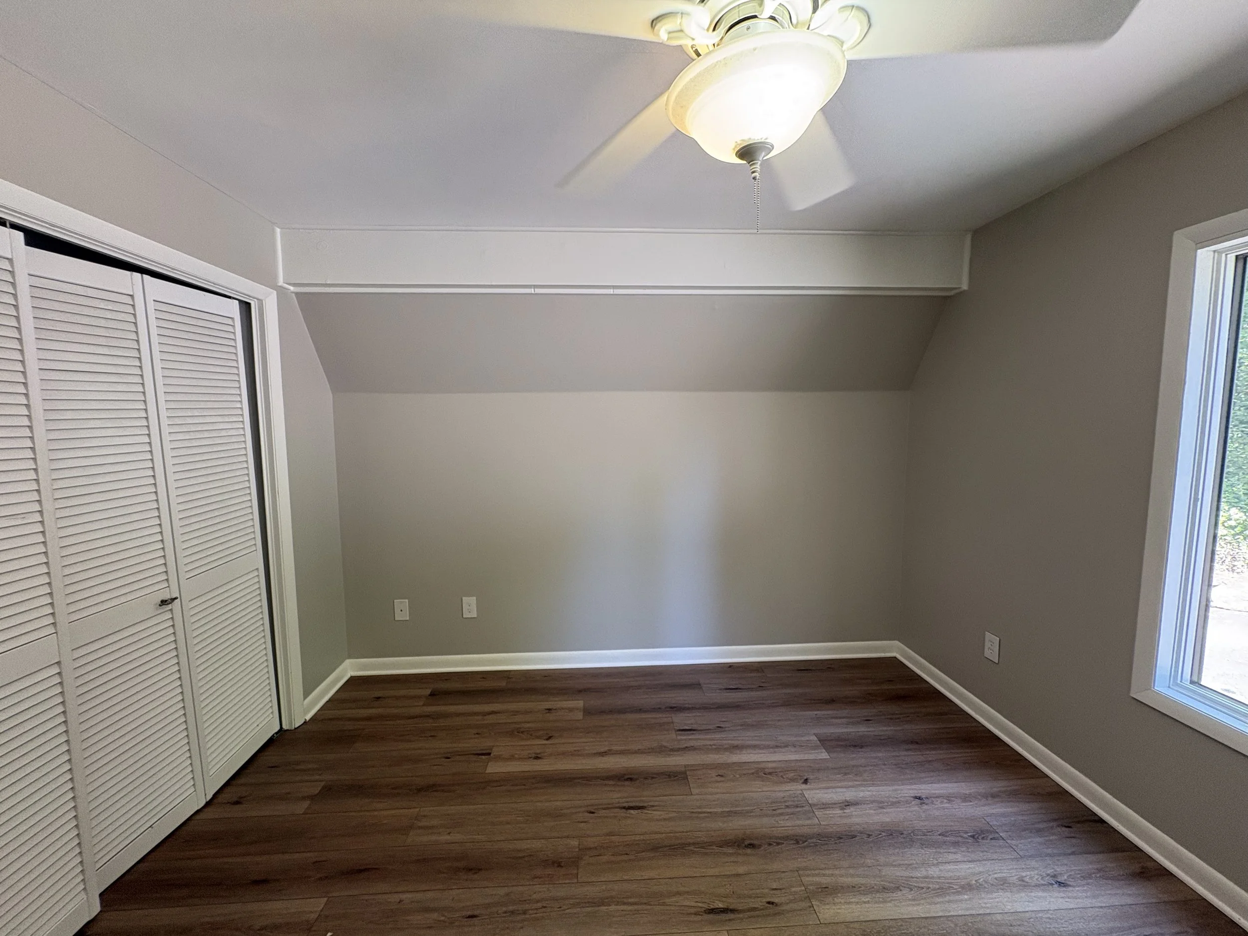 Empty room with white walls, hardwood floor, a ceiling fan with a light, and a window with a view outside.