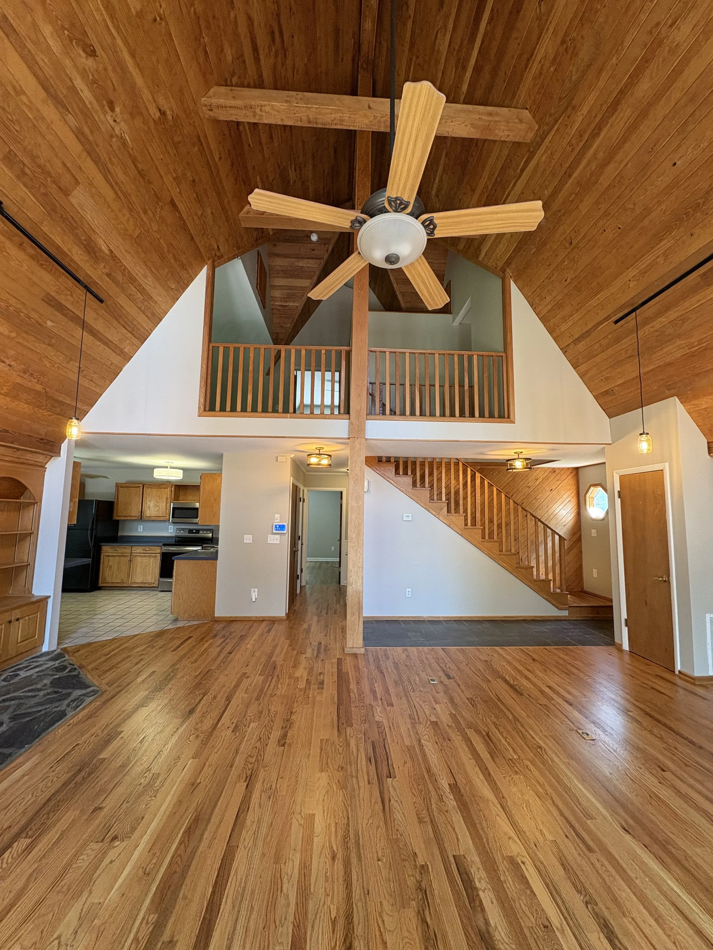 Interior view of a house with wooden floors, vaulted wooden ceiling, wooden stairs, and a loft area with railing. Part of kitchen visible with wooden cabinets. Ceiling fan hanging from the ceiling, pendant lights, and a small window near door.