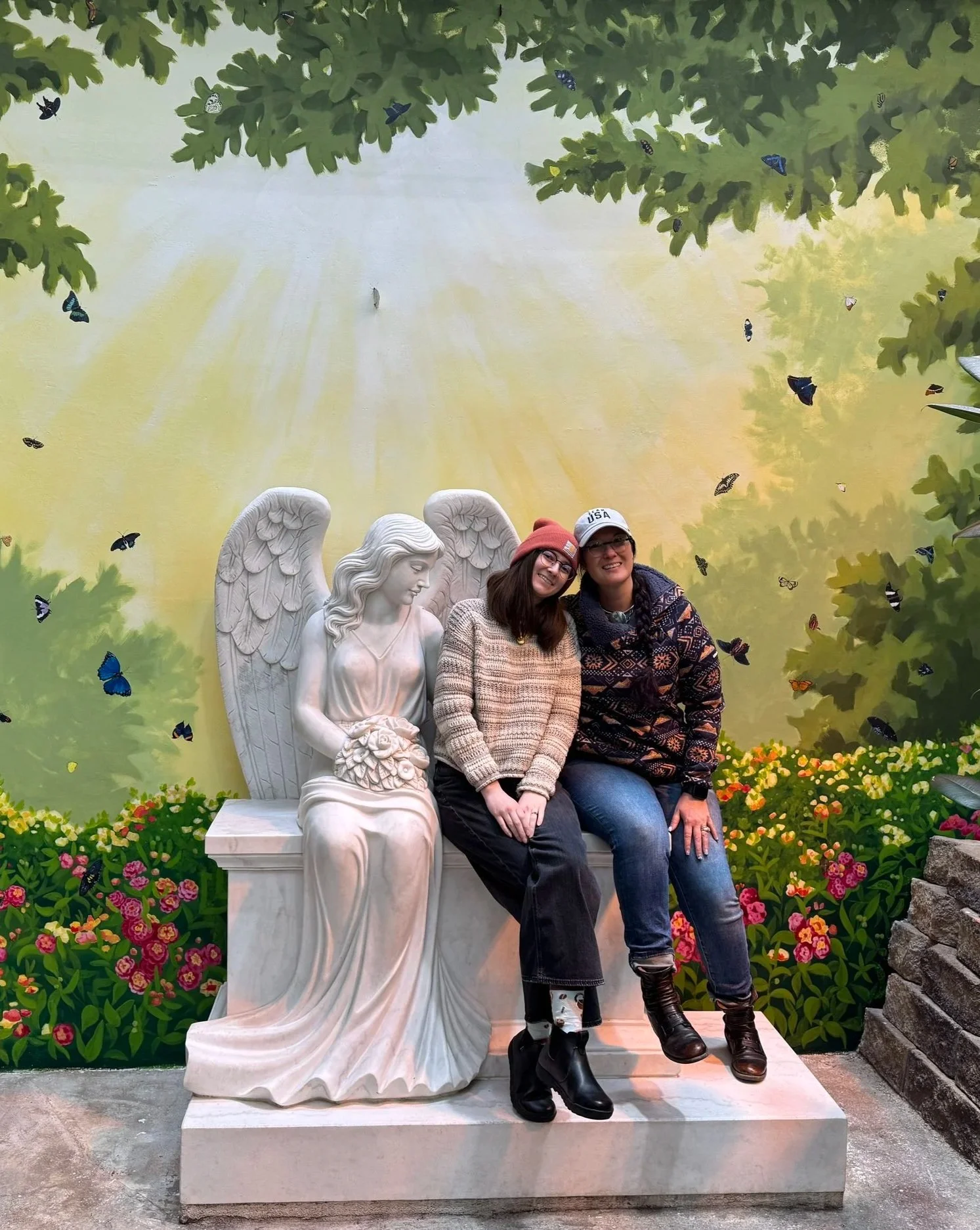 Two people sitting on a white statue of an angel with wings, surrounded by vibrant flowers, in front of a painted backdrop of trees and butterflies, in a garden or park setting.