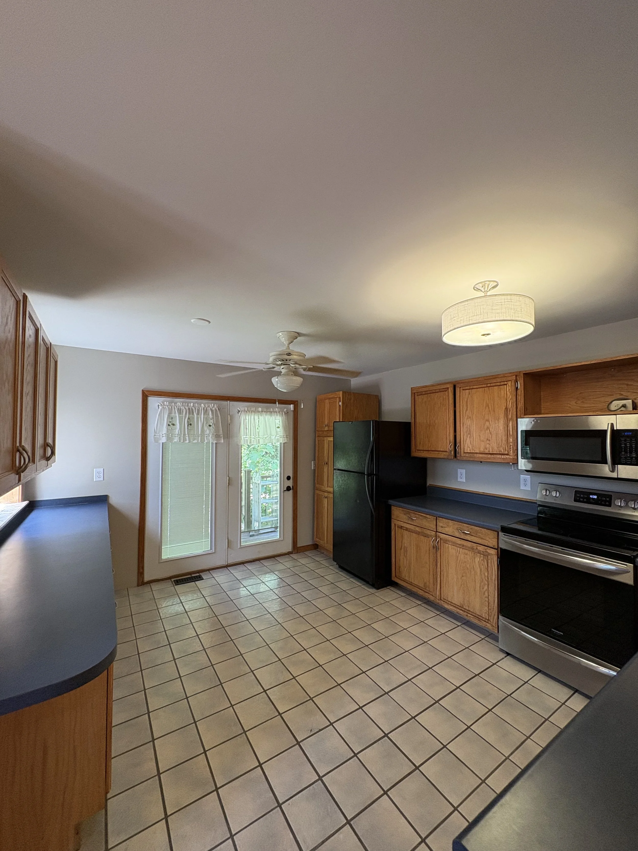 A kitchen with wooden cabinets, black countertops, and stainless steel appliances including a microwave and oven. White ceiling with a ceiling fan and a light fixture. There is a door with glass panes and curtains, leading outside, with a tiled floor