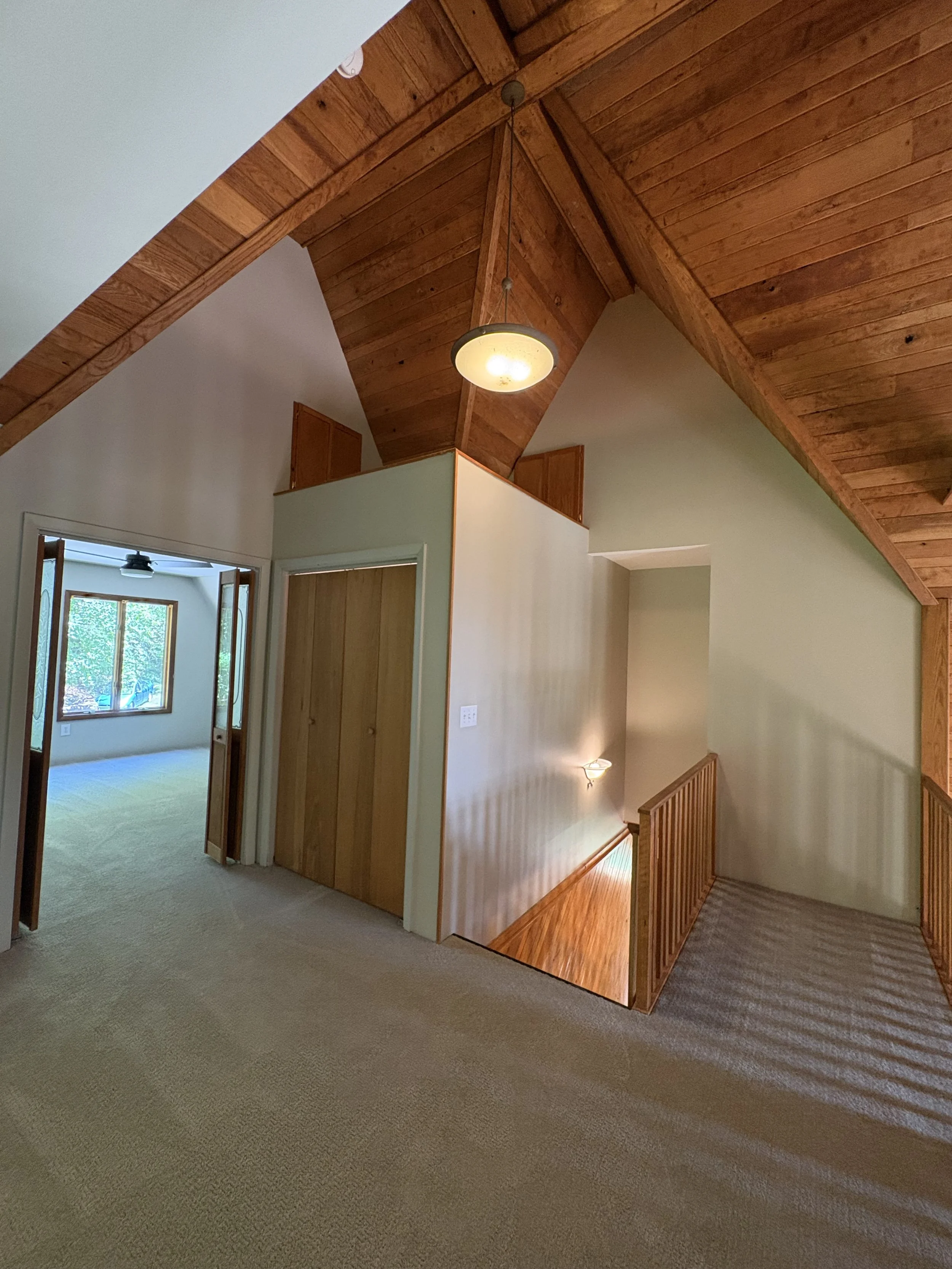Photo of an interior upper level of a house showcasing a wooden vaulted ceiling, a hanging light fixture, a staircase with wooden railing, and rooms with open doors leading to a sunny room with windows.