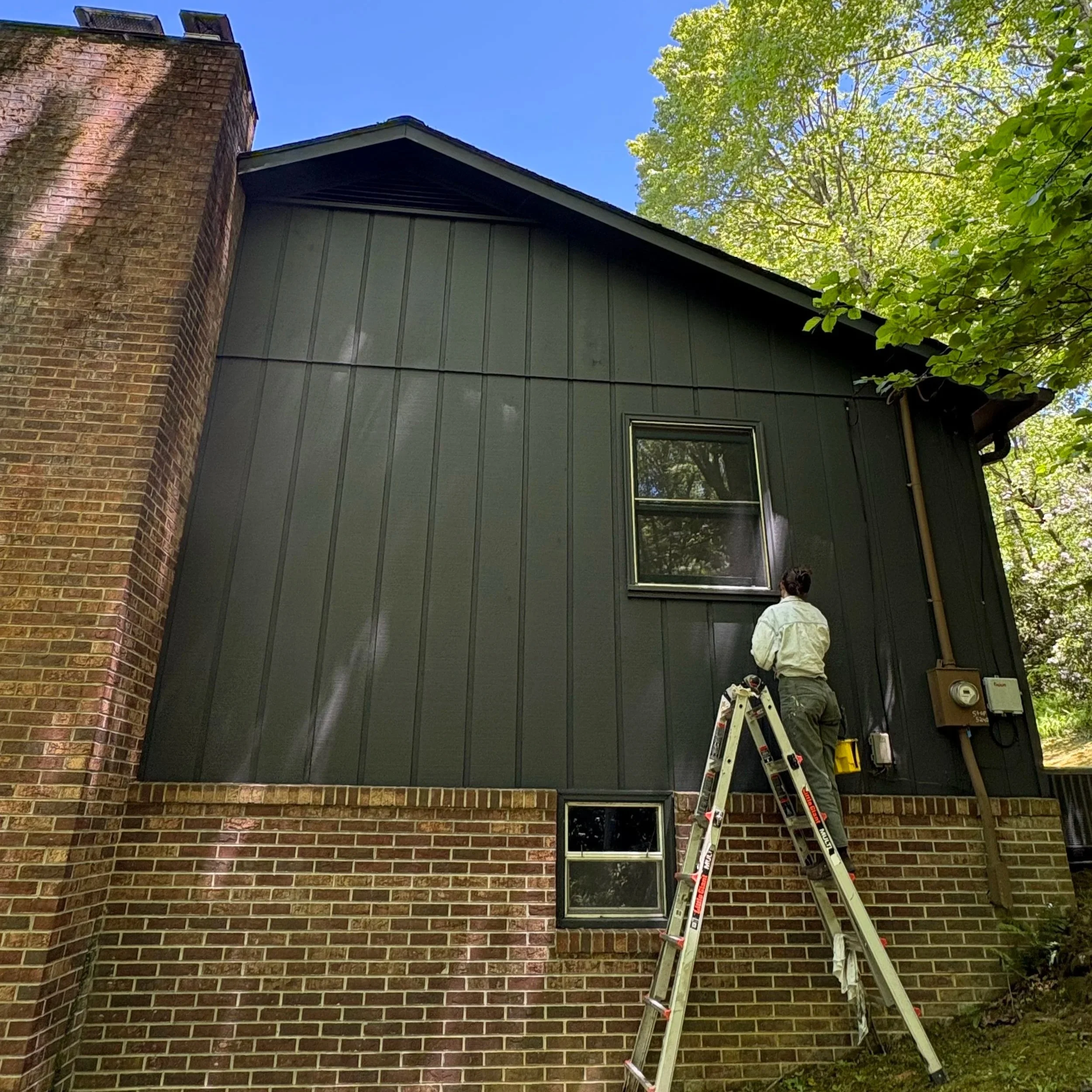 Man on a ladder working on the exterior house wall, with a brick chimney and window, surrounded by green trees and blue sky.