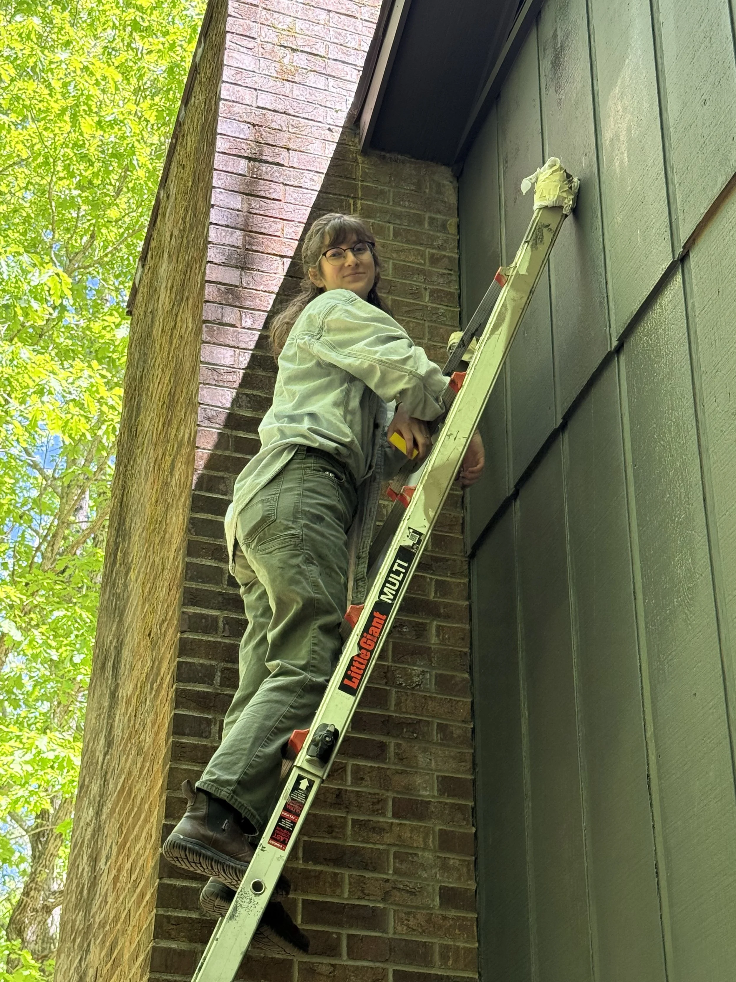 A young woman with glasses, long hair, and casual attire standing on a ladder against a brick wall and siding, working on the house exterior, surrounded by green trees.