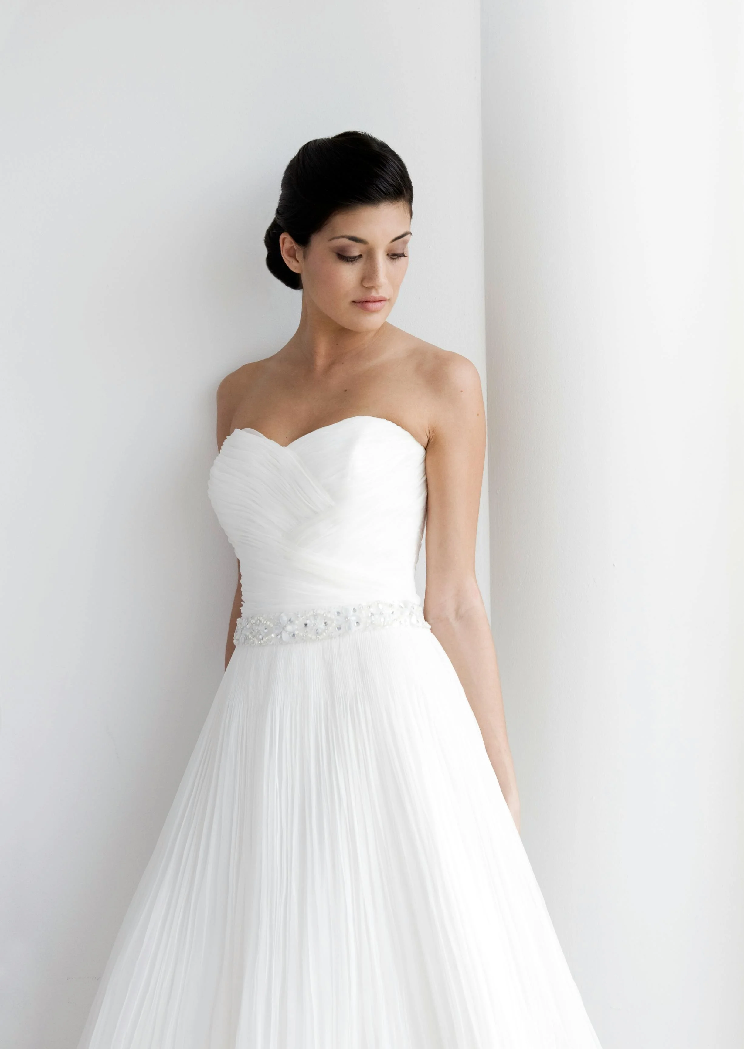 Bride with dark hair in a bun, standing in a corner of a room, wearing a simple strapless wedding dress and looking down.