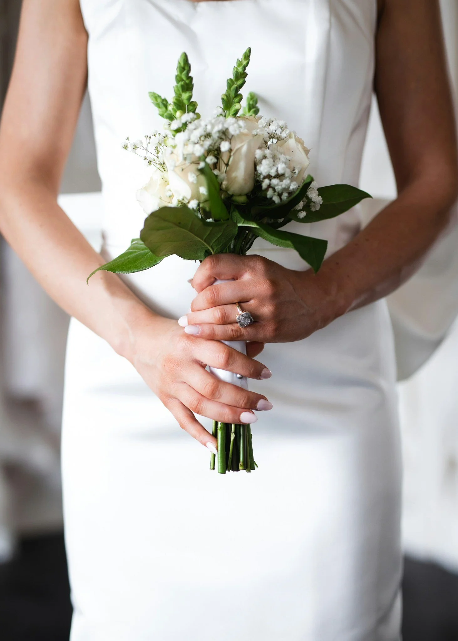 A bride in a sleek white wedding dress, with her hands in front of her, holding a simple bouquet of greenery and white roses.