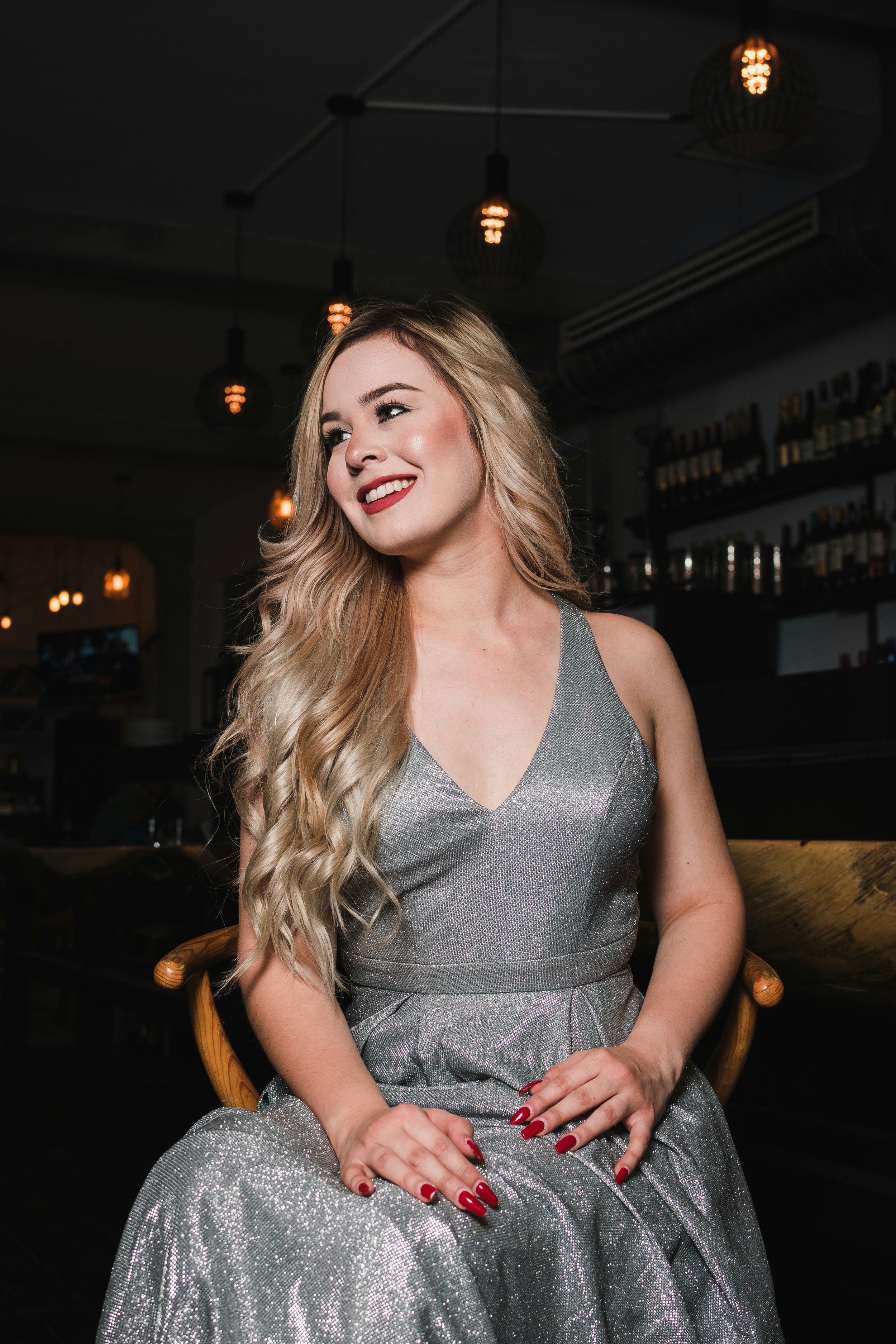 Young woman with long blonde hair sitting in a chair in a dark room, wearing a glittery v-neck silver dress.