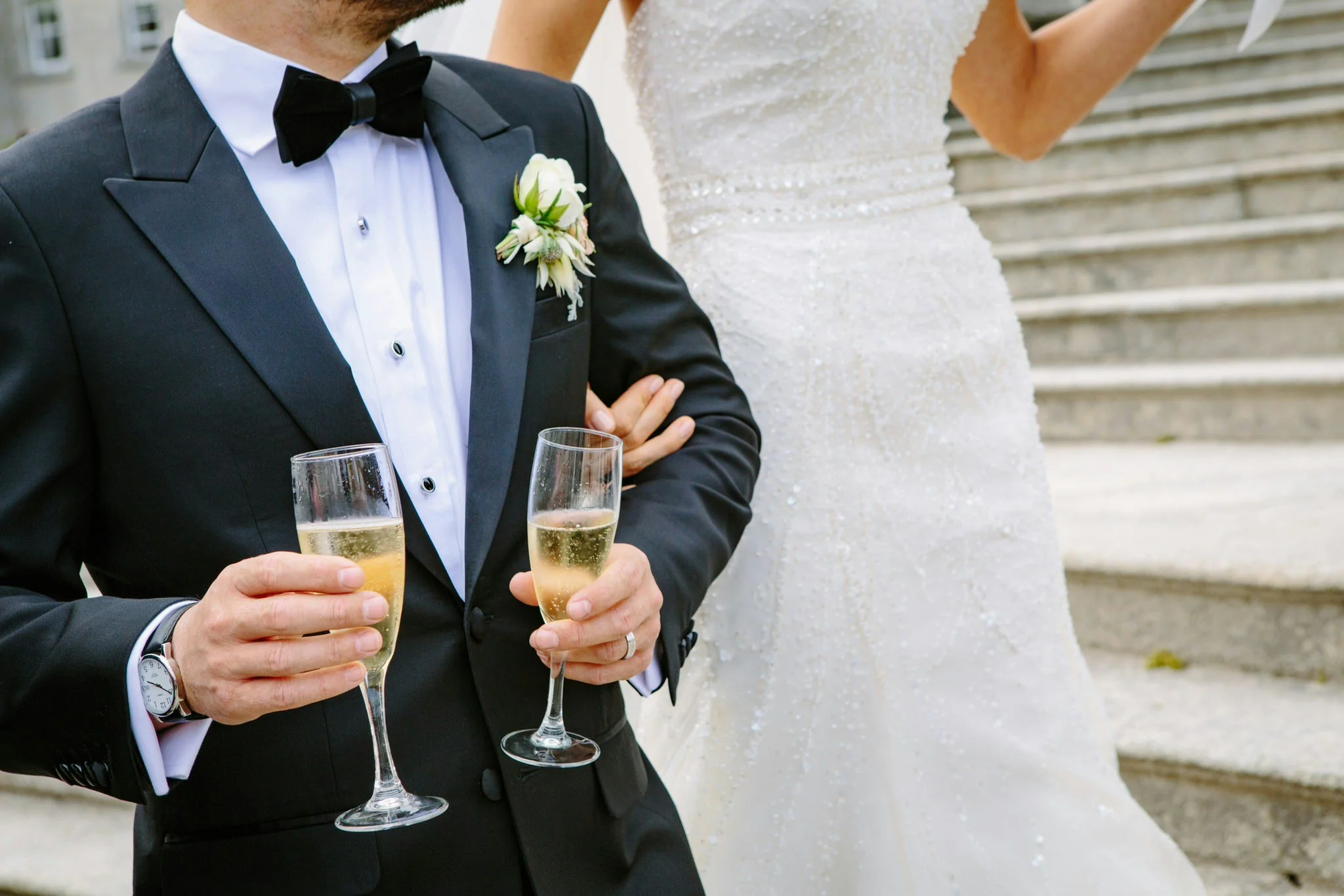 Close-up of a man in a tuxedo holding two glasses of champagne at a wedding, with a woman in a white wedding dress standing beside him. They are outdoors with steps in the background.