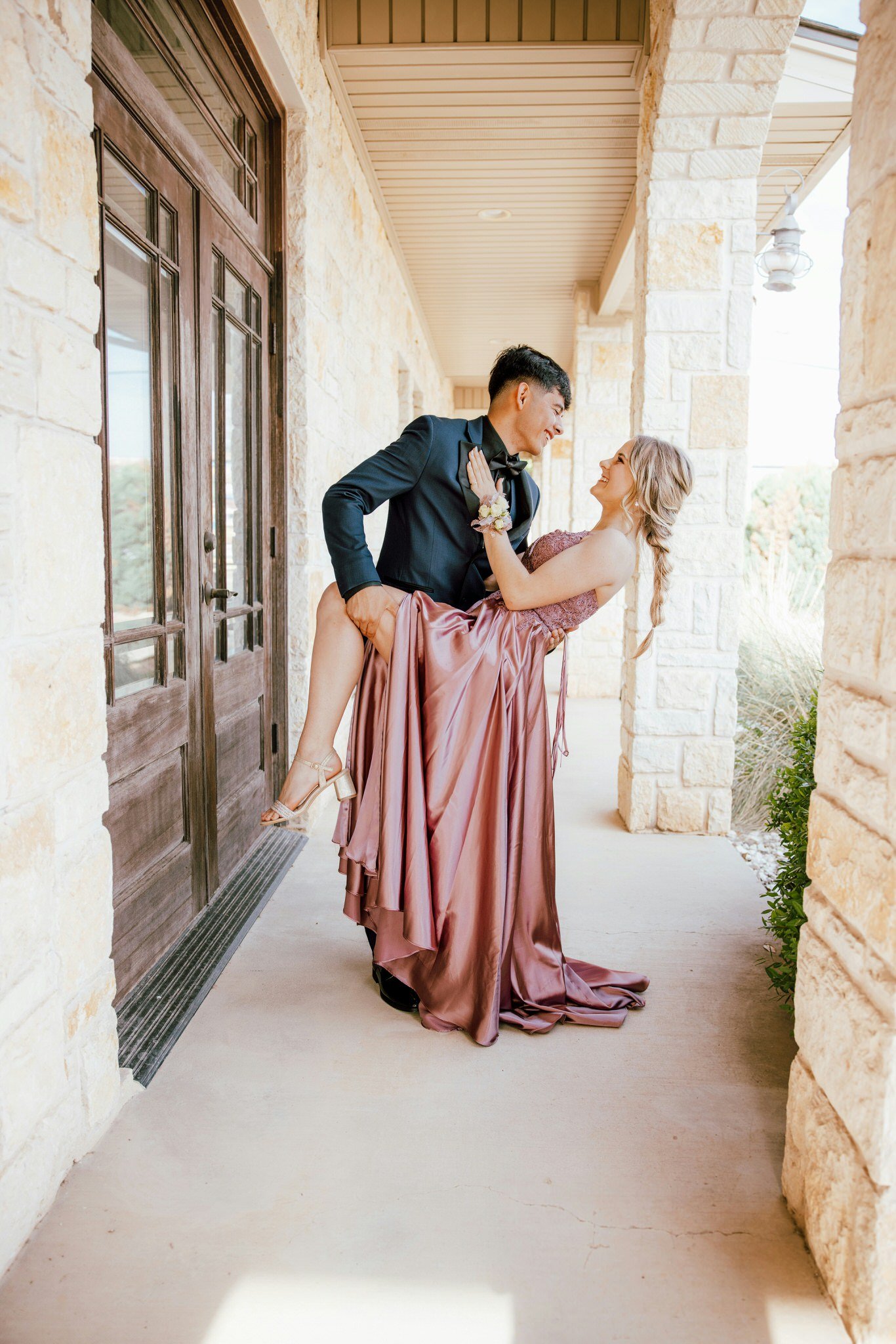 A couple dressed in formal attire, with the man in a black tuxedo and the woman in a pink satin gown, smiling at each other on a porch with stone walls and a wooden door.