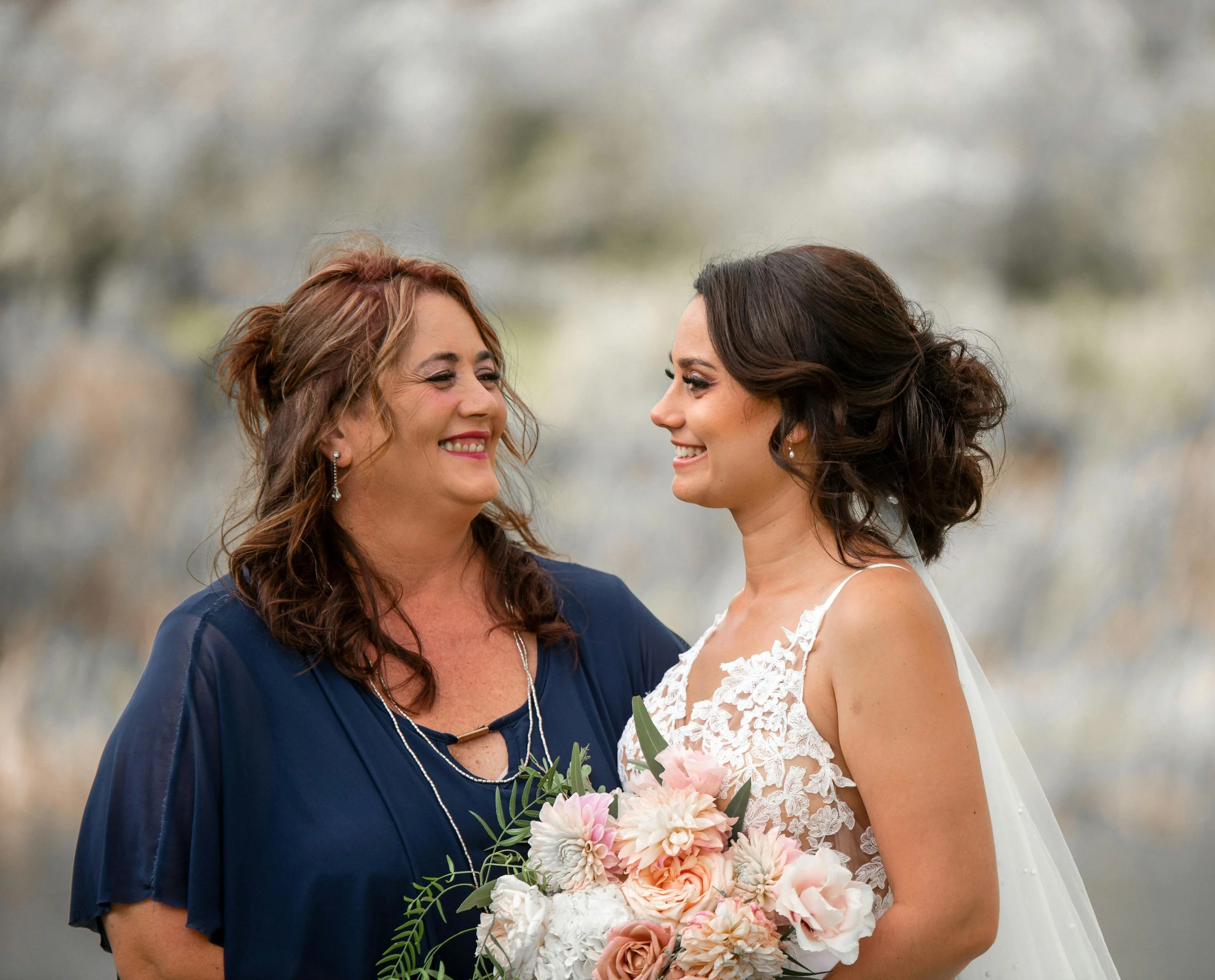 Middle-aged woman with brown hair, wearing a navy blue formal dress, looking at a younger woman in a wedding dress holding a bouquet, with both women smiling towards each other.