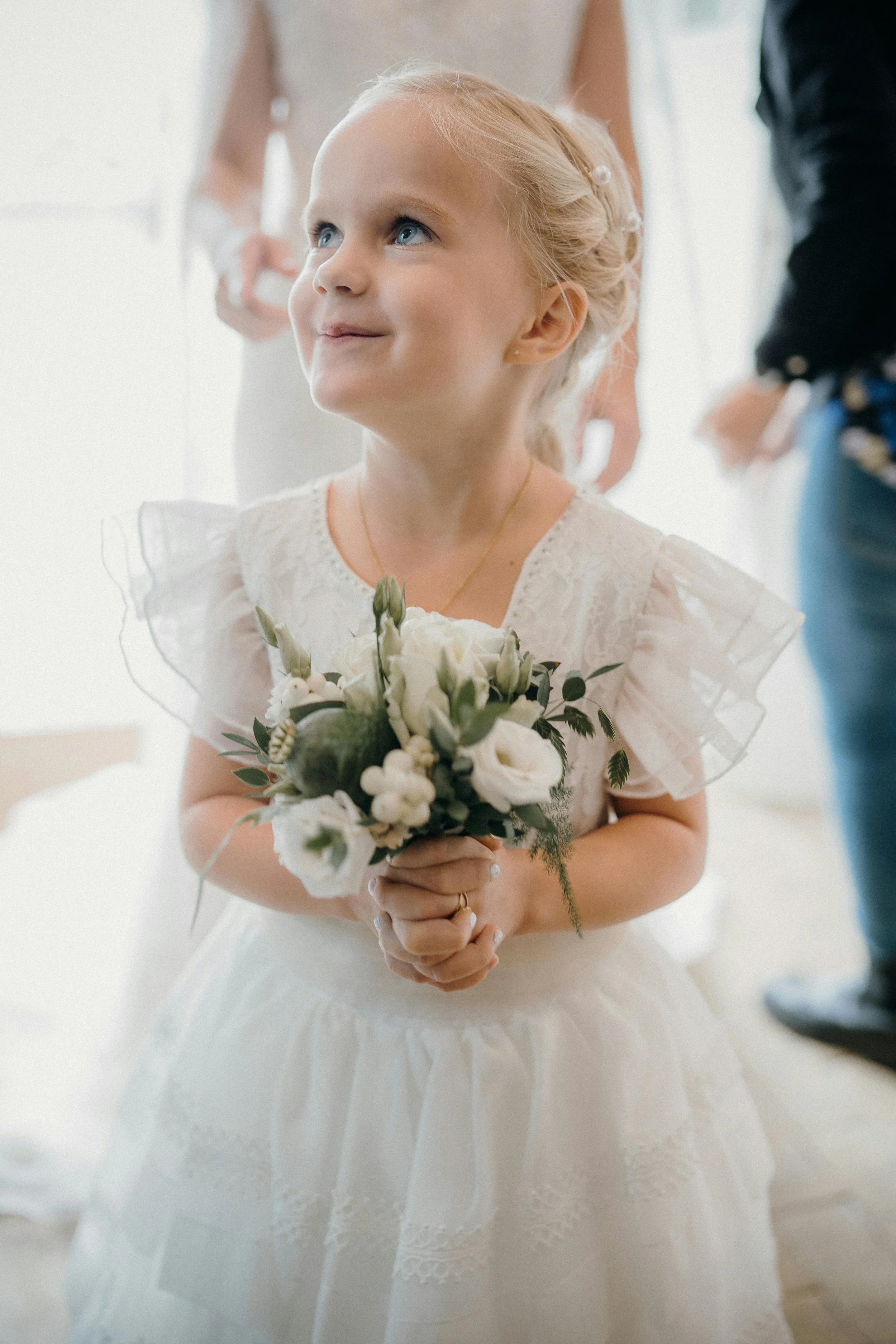 Young girl with blonde hair wearing a white dress with flutter sleeves and carrying a small floral bouquet.