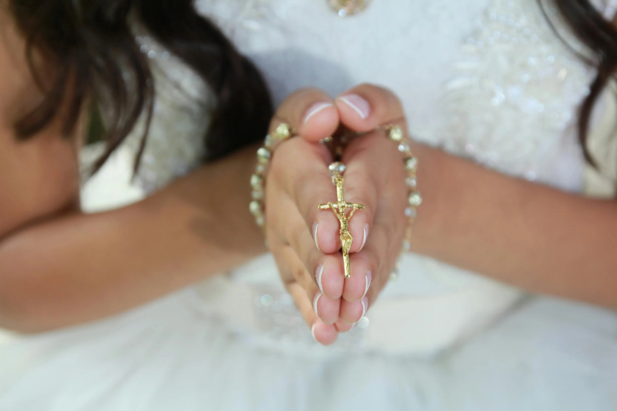 Close-up of a young girl holding a gold rosary in her hands and wearing a white dress.