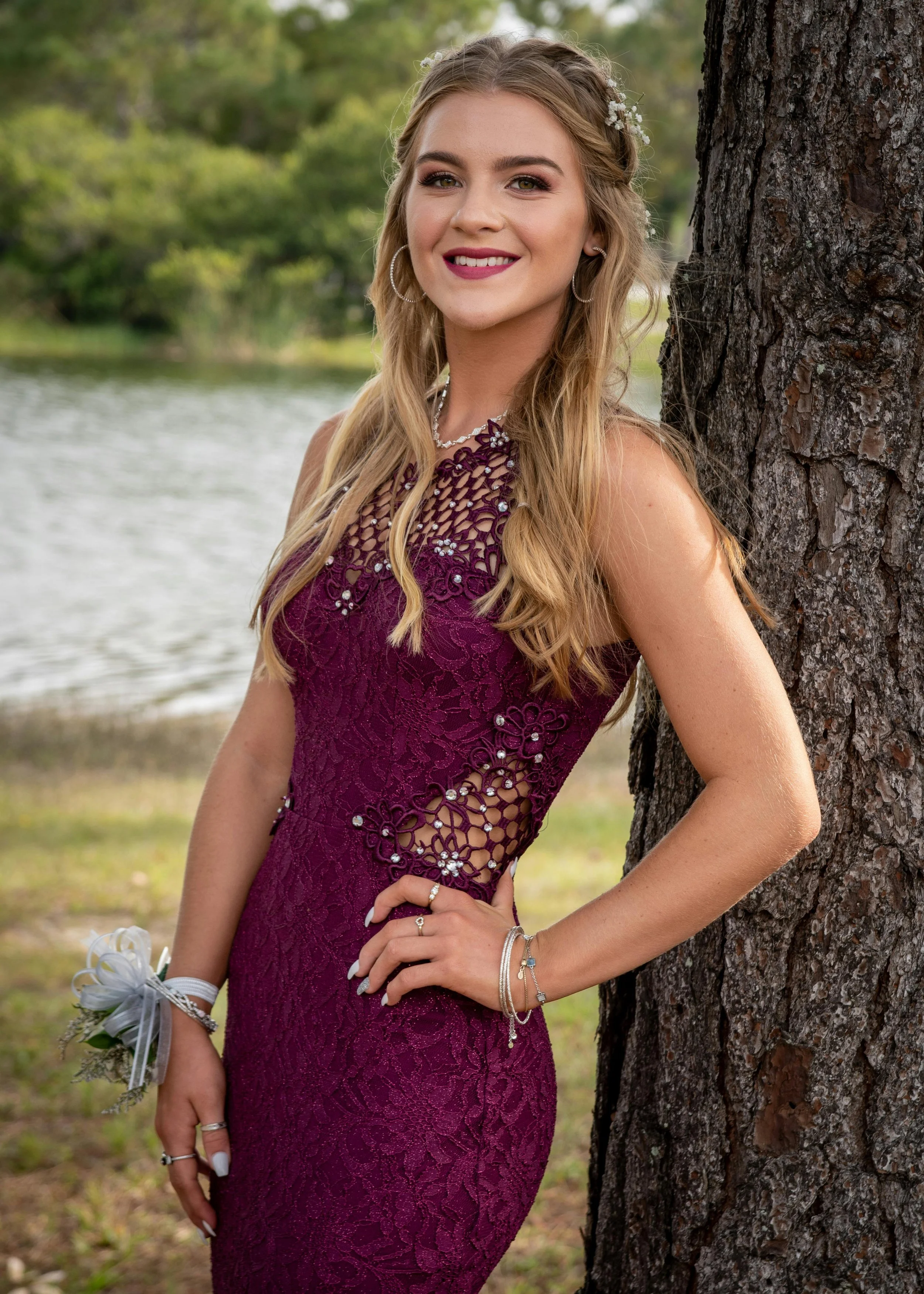 Teenage girl with blonde hair, wearing a dark pink lacy dress, leaning against a tree.