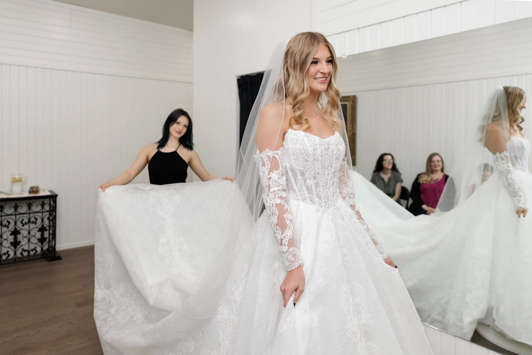 Young woman with curled blonde hair wearing an a-line white wedding gown and veil, in a room with a large mirror, while a woman wearing a black outfit is in the background fluffing the long train of the dress and other onlookers are smiling.