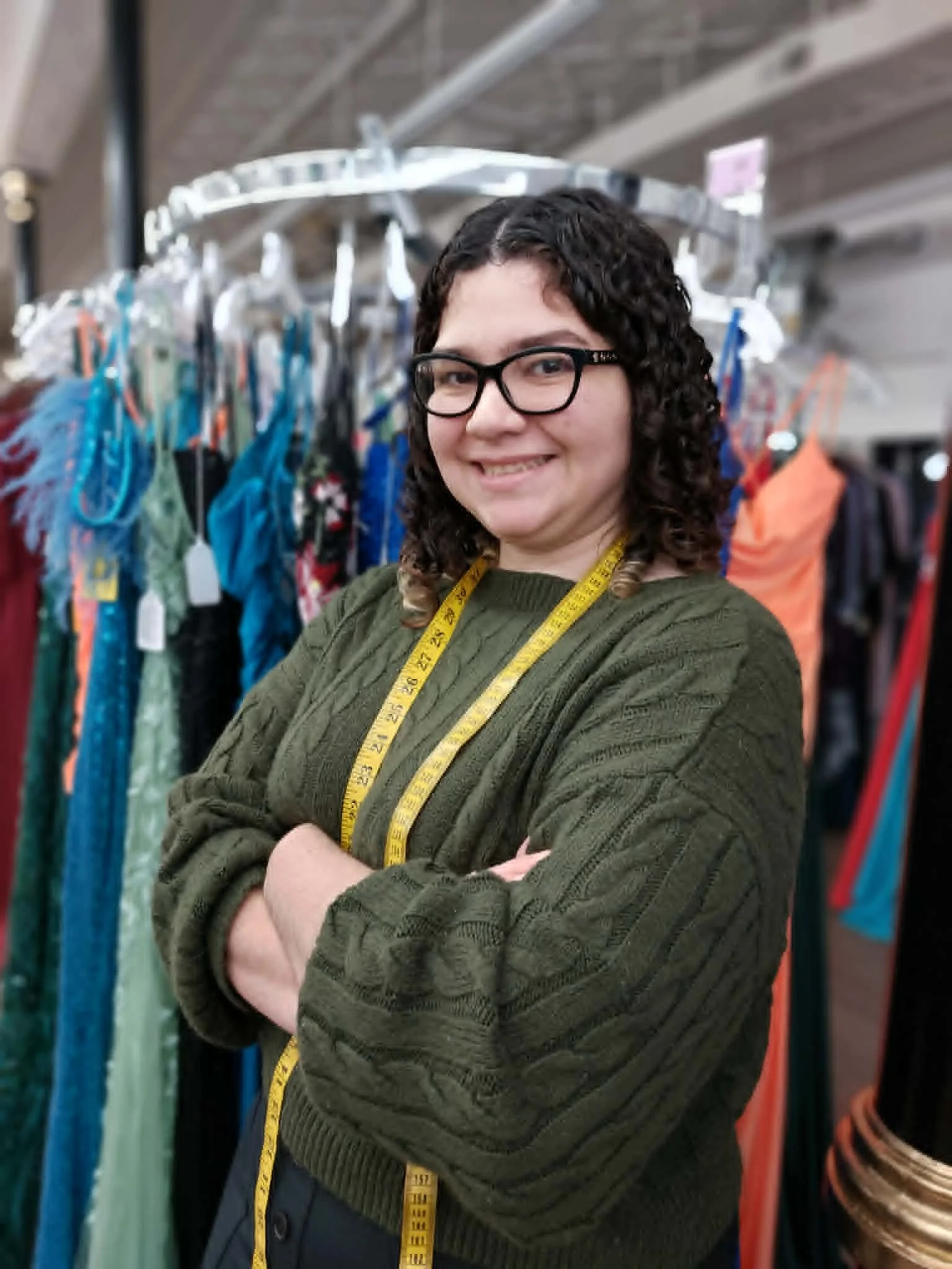 A woman with dark curly hair and glasses smiling and standing with her arms crossed in front of a clothing rack with various garments, including dresses and shirts, in a store.