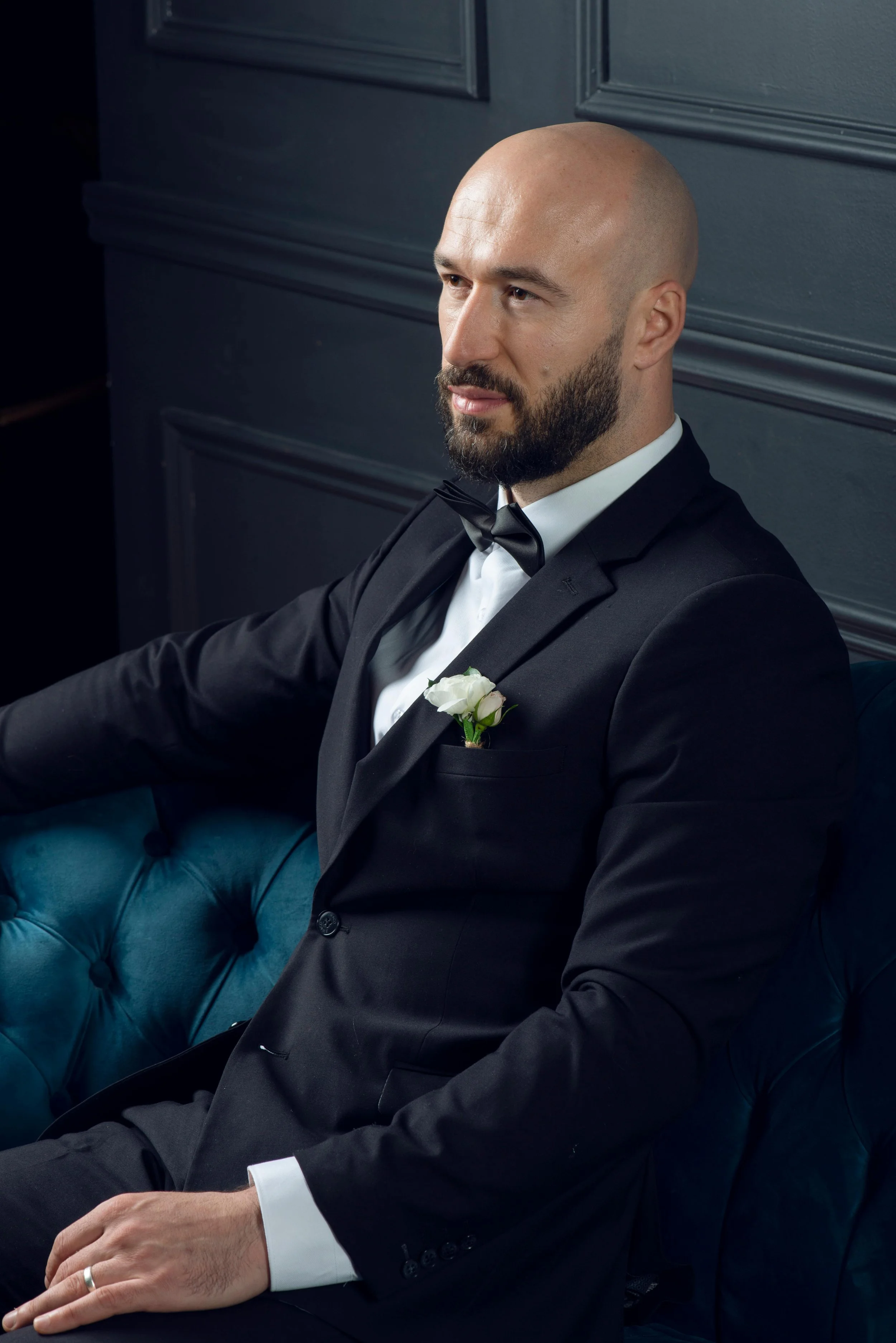 A man with a bald head and dark bear sitting on a teal tufted chair, wearing a black tuxedo, white shirt, and black bowtie, with a white flower in the front pocket.
