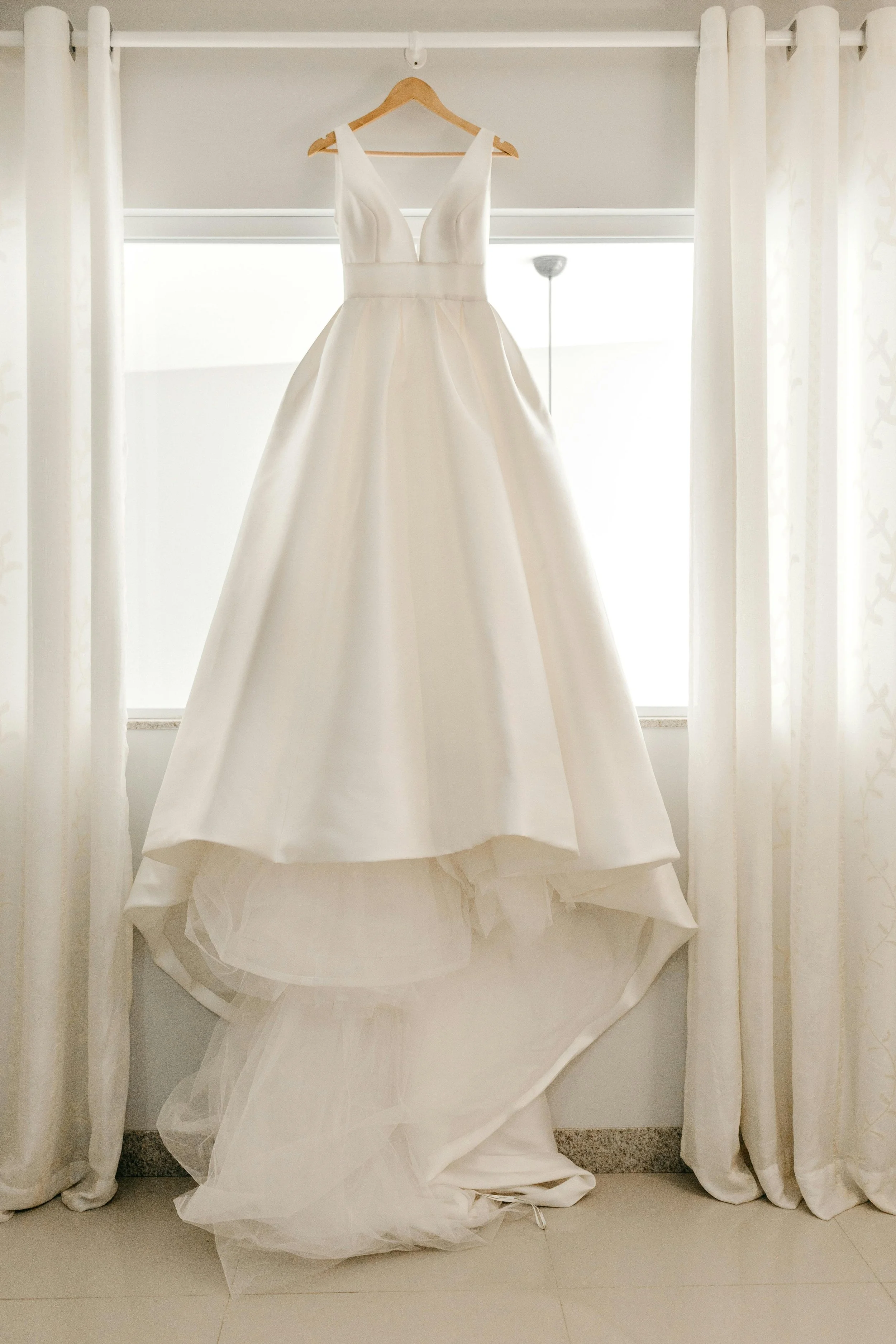 A white wedding dress hanging on a wooden hanger in front of a window with white curtains.