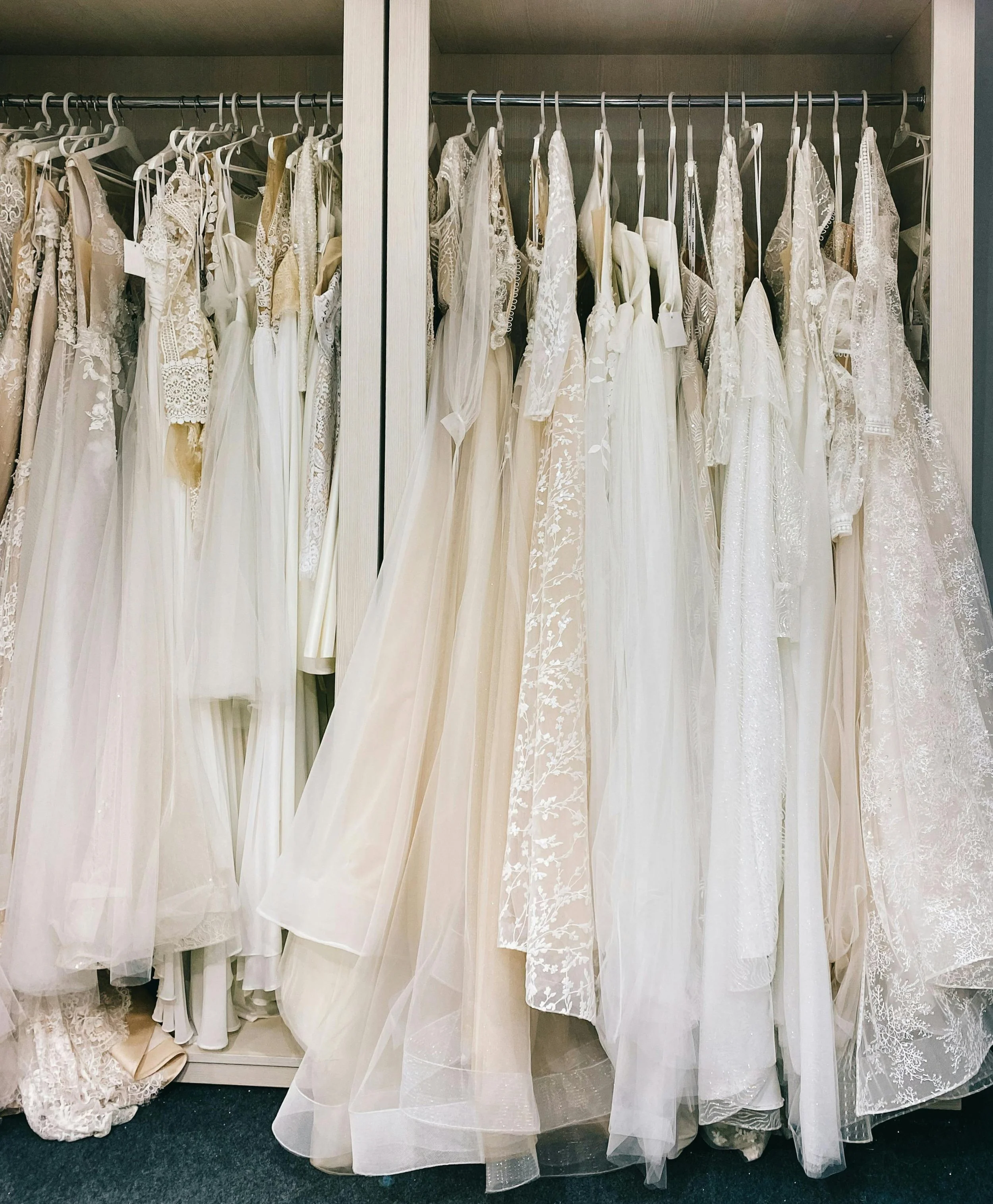 A closet with a metal rod holding numerous wedding dresses on hangers, featuring lace, tulle, and satin fabric in shades of white and ivory.