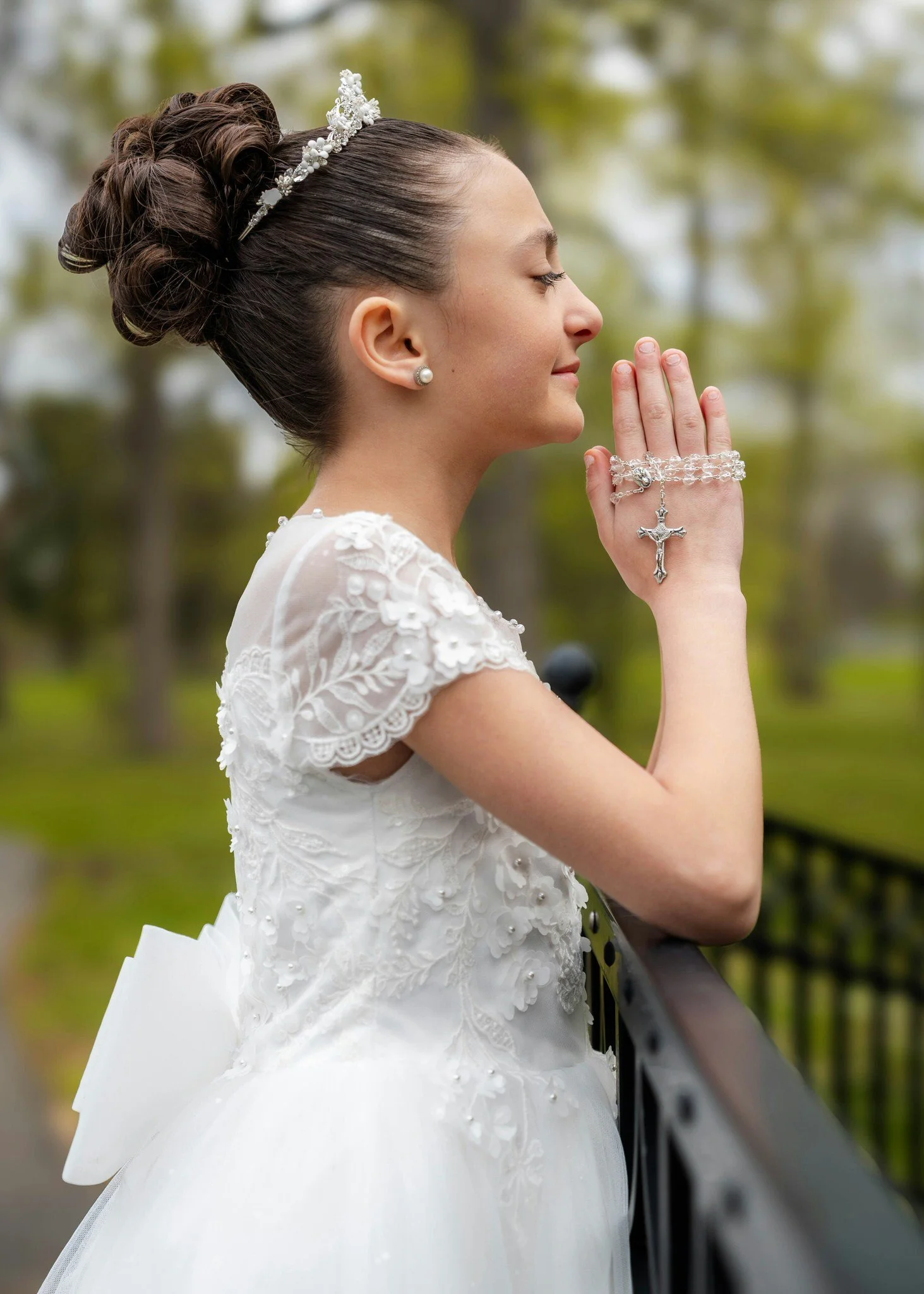 Young girl wearing a white first communion dress, with her hands folded in prayer, and a rosary wrapped around her hands, leaning against a black iron fence outdoors.