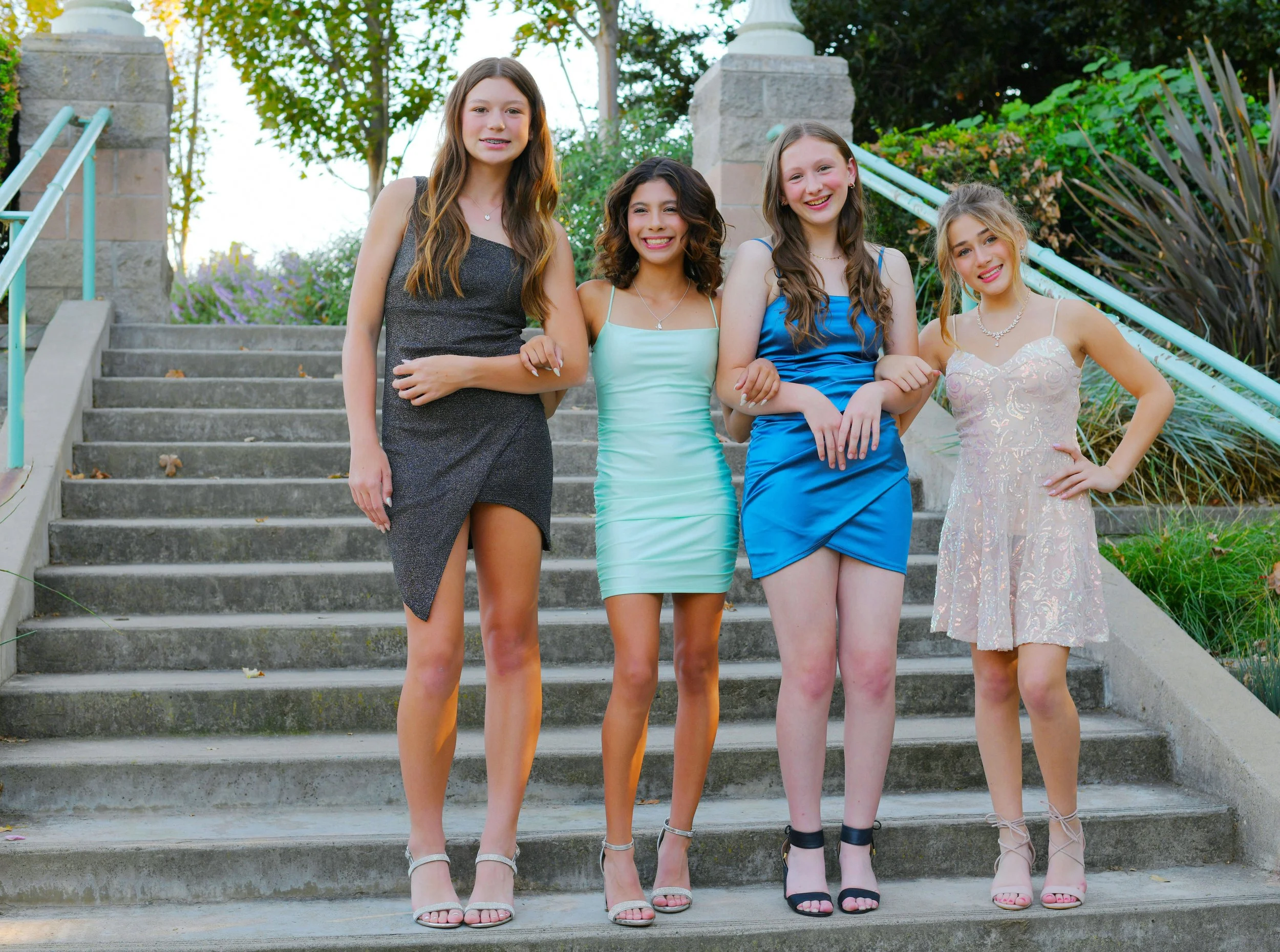 Four teen girls standing on concrete stairs while linking their arms together, dressed in short dresses in a variety of colors.