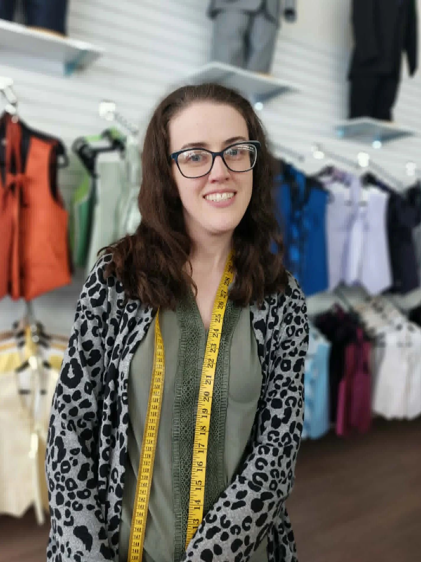 A woman with long brown hair and glasses smiling at the camera in a clothing store, wearing a green dress with a yellow measuring tape around her neck, a black and white leopard print cardigan, with clothing racks and shelves in the background.