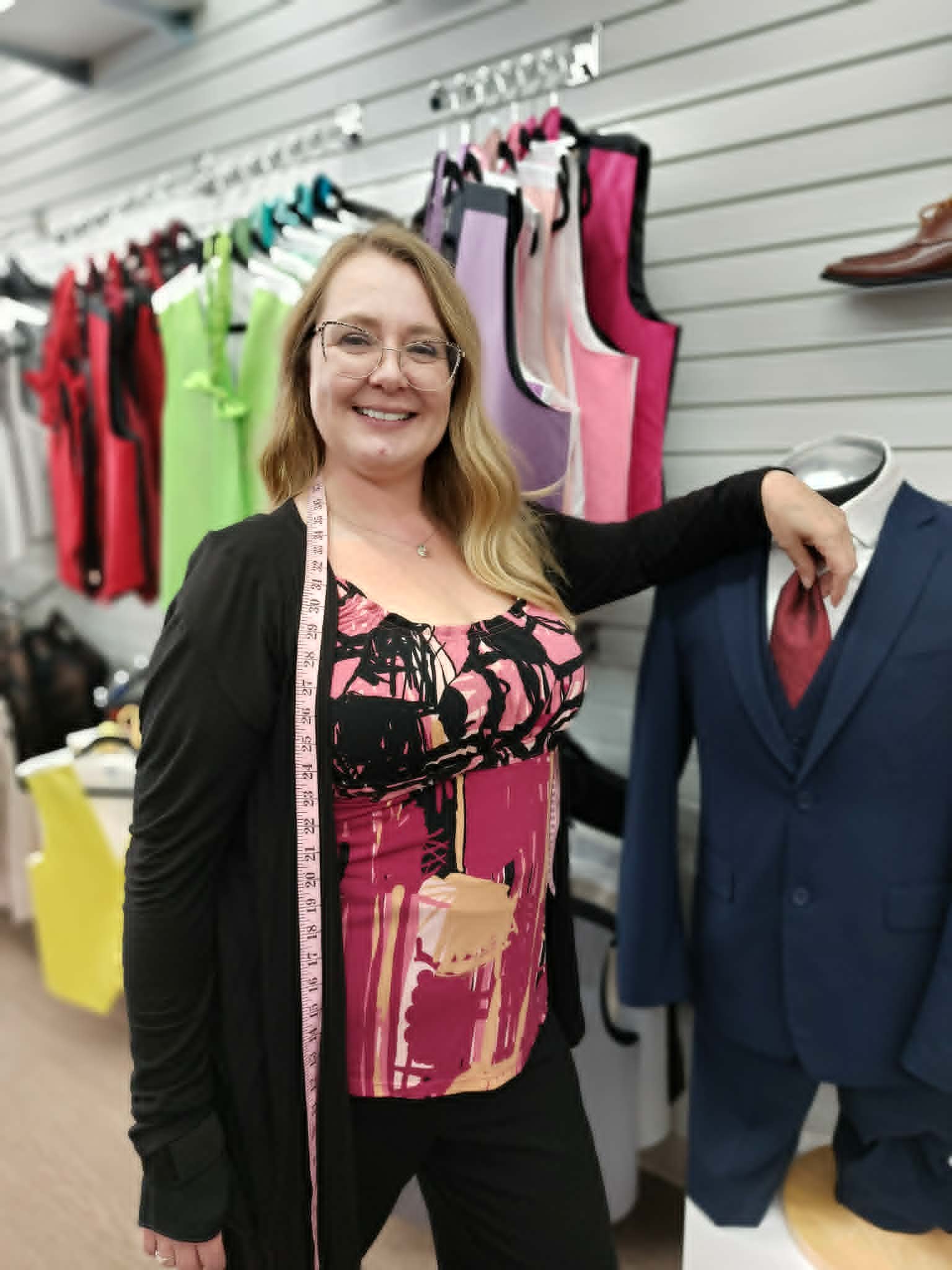 A woman smiling at a clothing store, standing next to a mannequin dressed in a navy suit with a white shirt and red tie. Behind her, colorful clothing items are hanging on a wall.