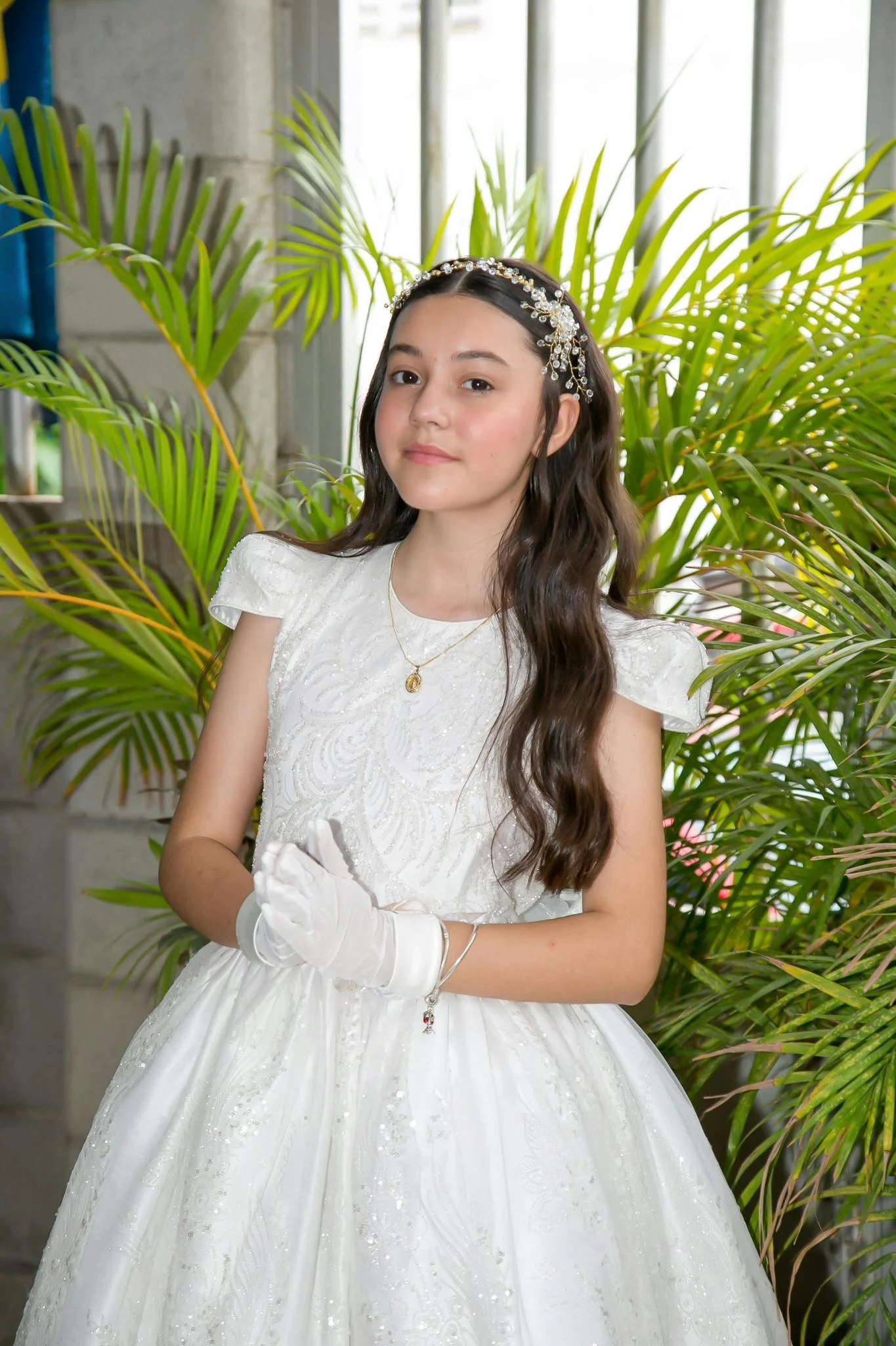 Young girl with long dark hair, wearing a white first communion dress and white gloves, with her hands together in a prayer, with green tropical leaves behind her.