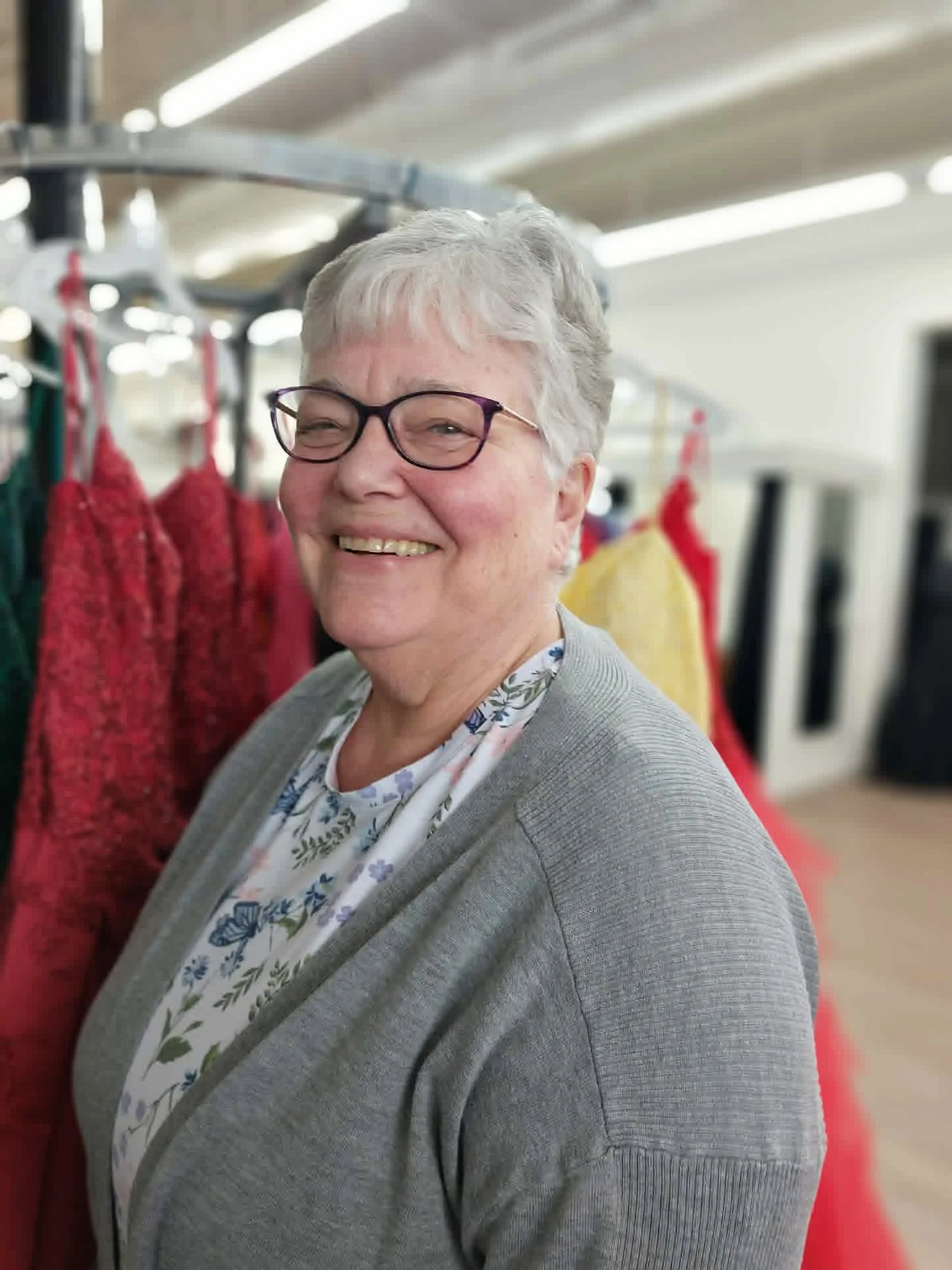 A smiling woman with short gray hair, glasses, and a floral blouse, standing in a store aisle with red and yellow clothing on racks.