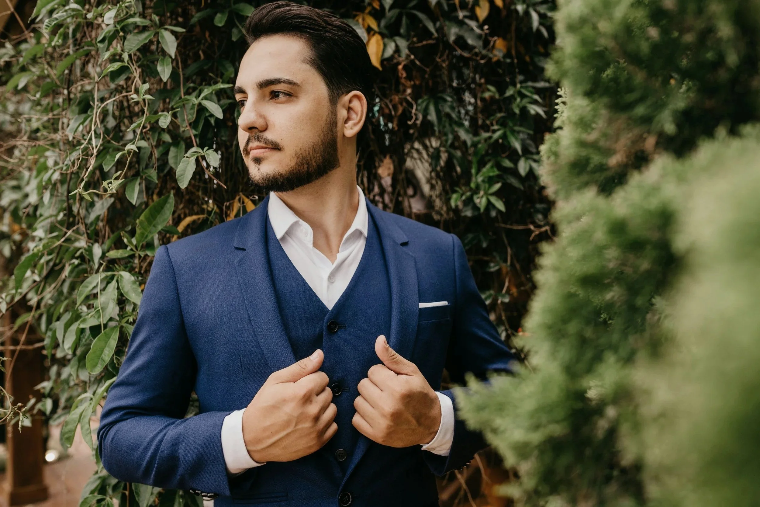 Man with brown hair and a beard, standing outdoors against green ivy and foliage, wearing a navy blue tuxedo and holding the lapels with both hands, while looking away to the side.