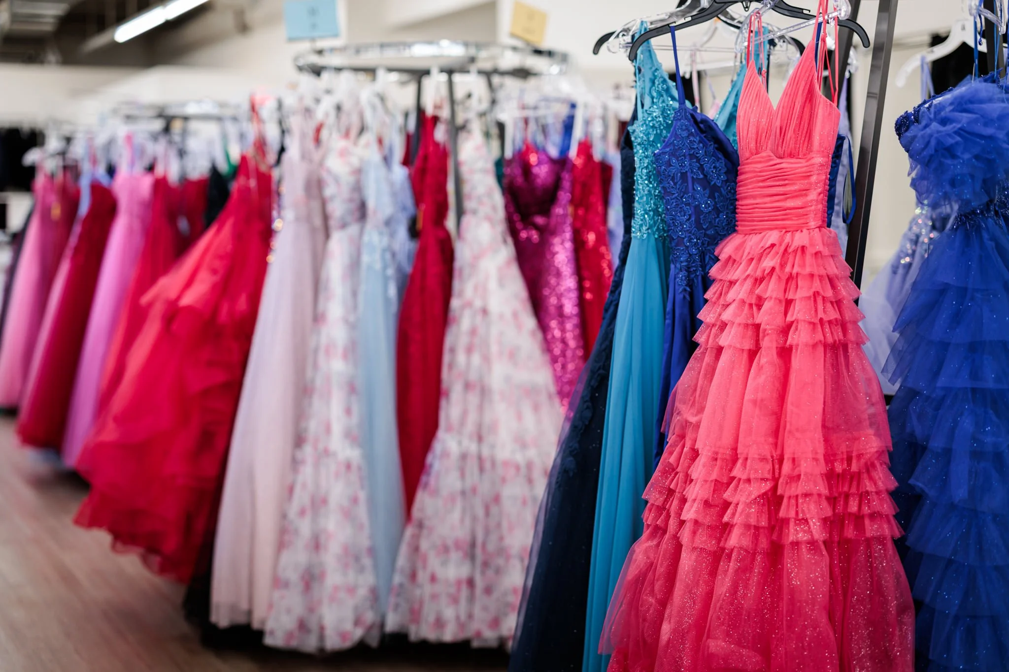 Colorful prom dresses in coral, blue, teal, and purple all hanging form a rack in a dress store, with other dresses blurred in the background.