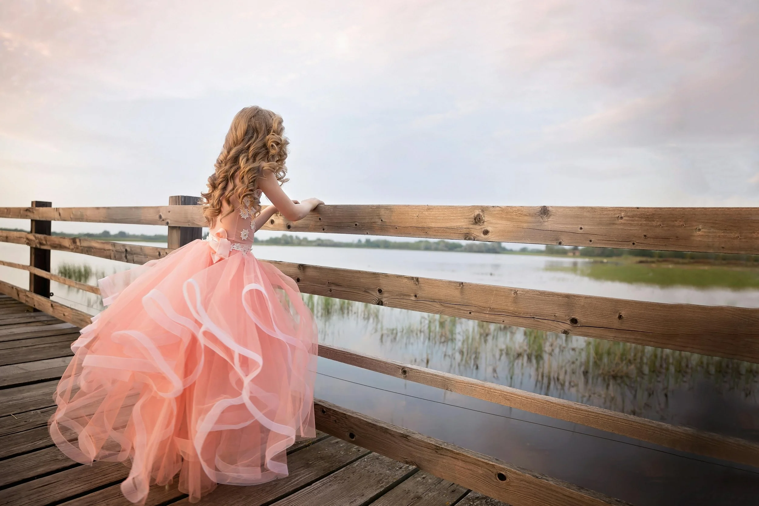Teen girl with curly hair wearing a poofy tulle prom dress, standing on a wooden bridge, looking out towards a lake.