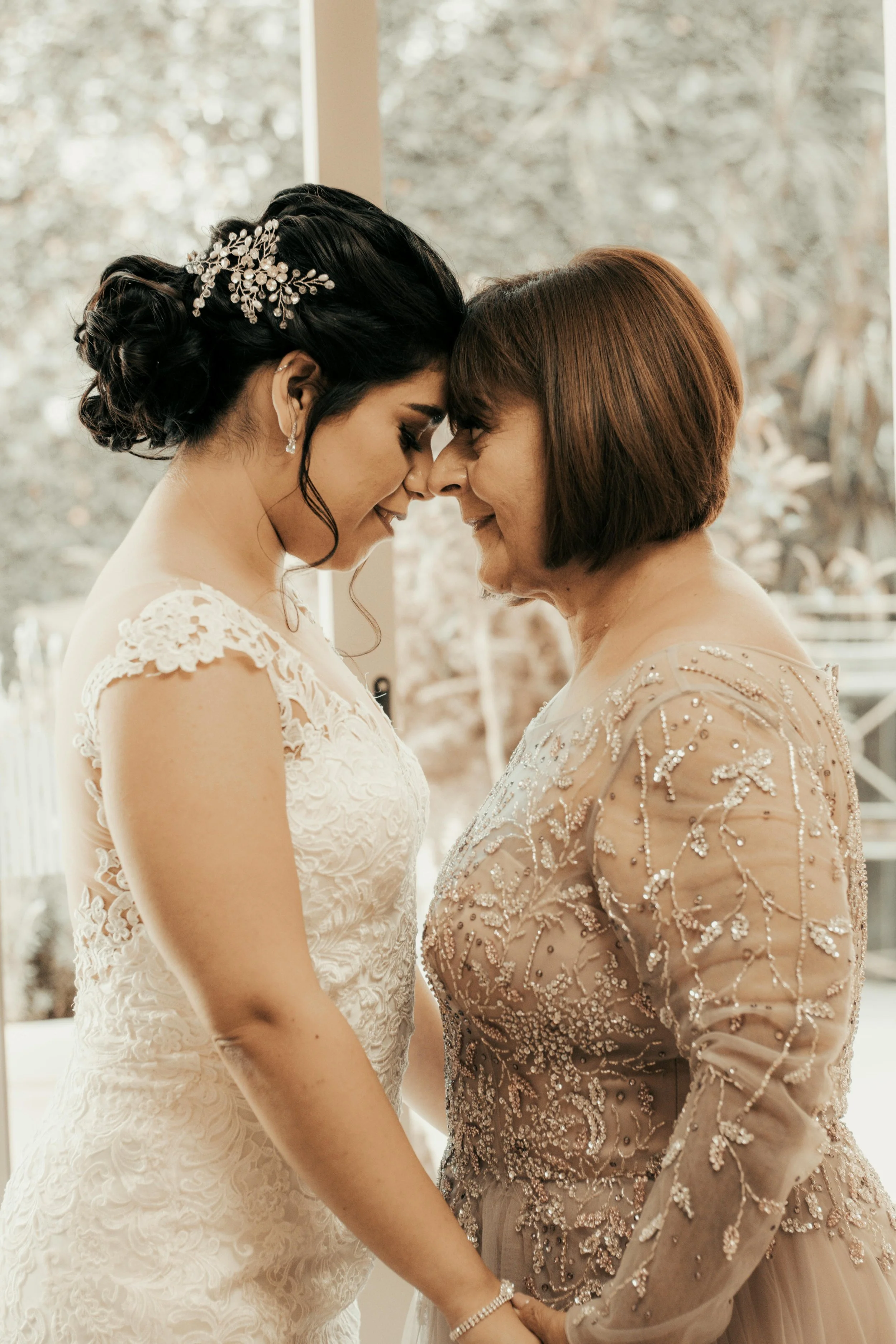 A bride with black hair in an updo leaning forehead to forehead with an older woman wearing a champagne gold embellished dress.