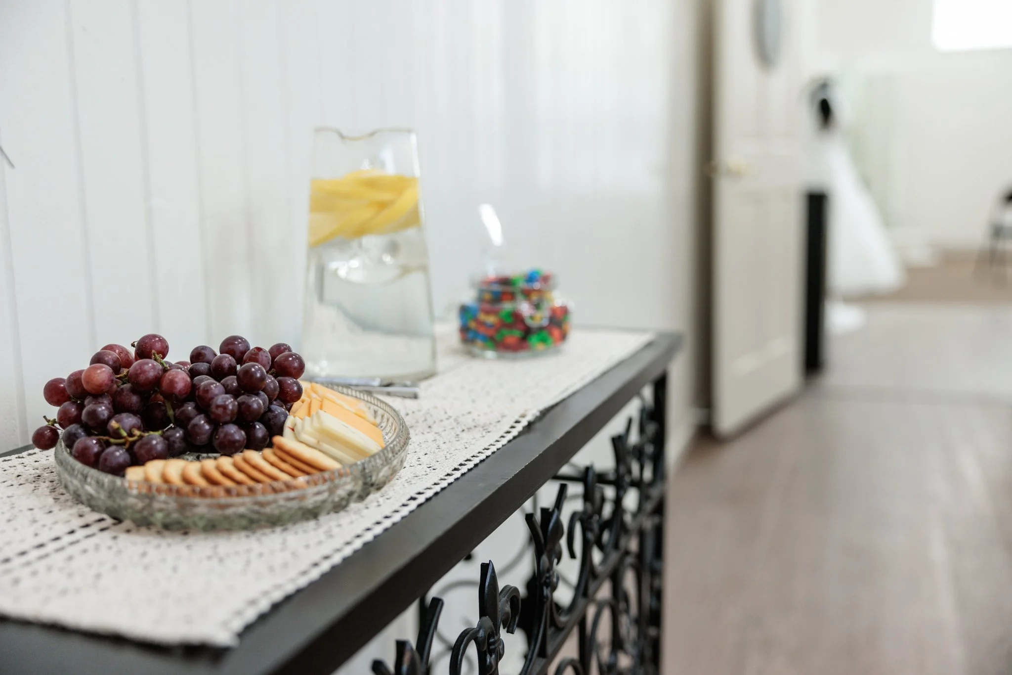 Small black console table with a tray of cheese, crackers, and red grapes sitting next to a pitcher of lemon water and a clear jar of candy. In the background is a room with a bridal gown on a mannequin.