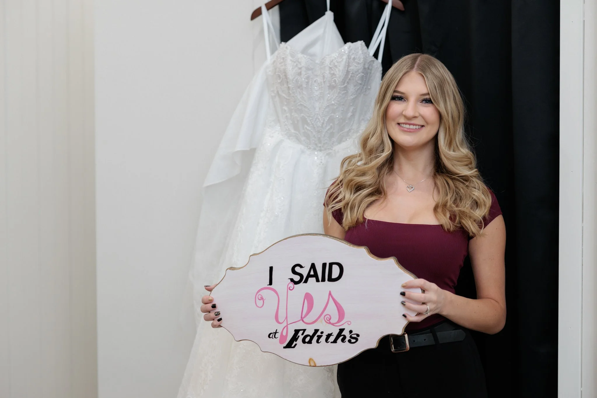 Young woman with a burgundy top and medium length blonde hair standing in front of a wedding dress, holding a sign that reads, "I said Yes at Edith's."