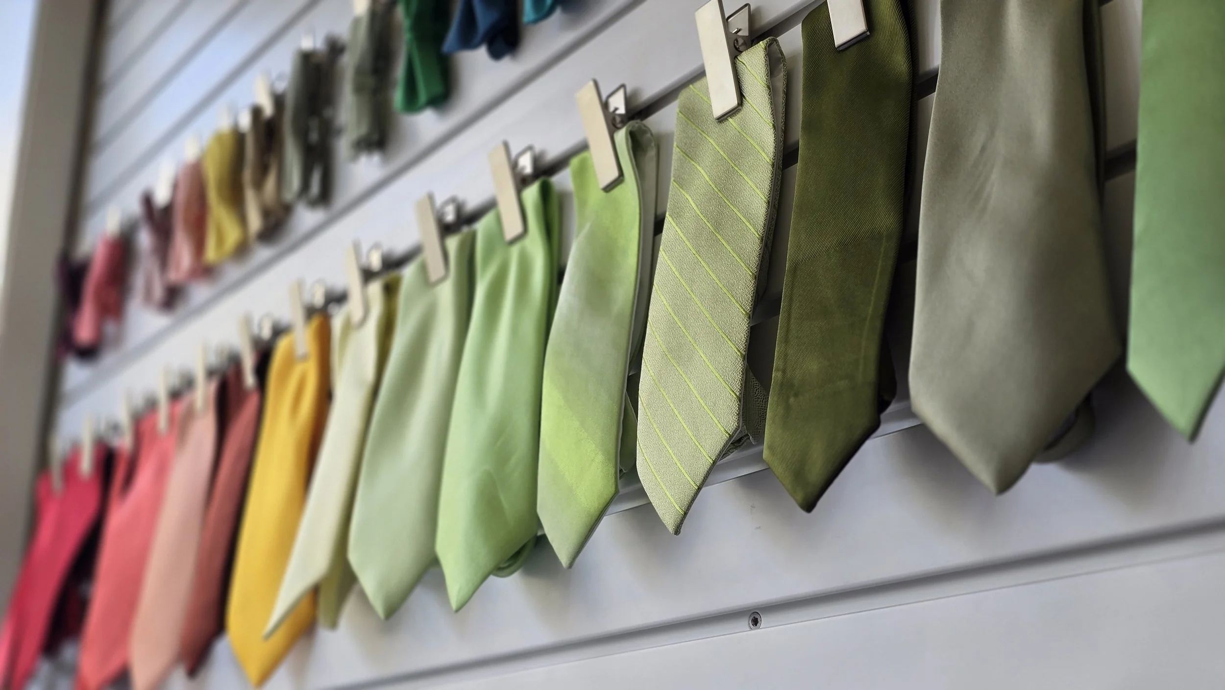 Display of colorful neckties hanging on a slatwall rack in various shades of pink, yellow, green, and gray.